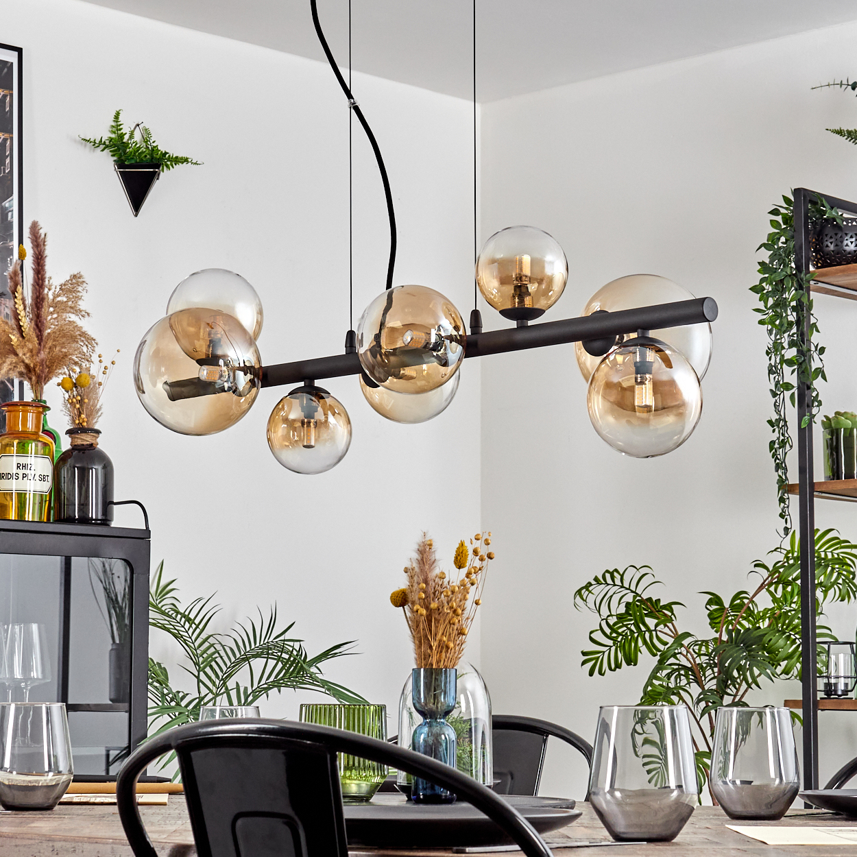 Ein Esszimmer mit einem schwarzen Kronleuchter mit Glaskugeln, einem Holztisch und Pflanzen. Gläser und Stühle sind ebenfalls vorhanden.