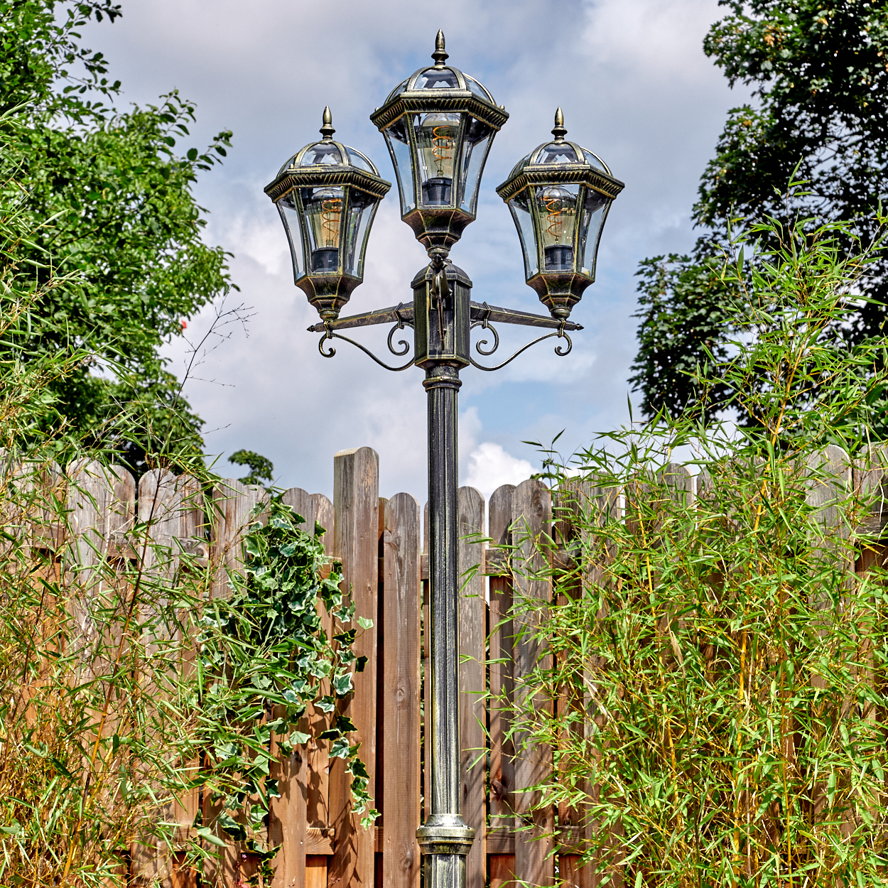 Eine dekorative Laterne mit drei Lampen steht in einem Garten. Ein Holzzaun und grüne Pflanzen sind im Hintergrund. Der Himmel ist blau.