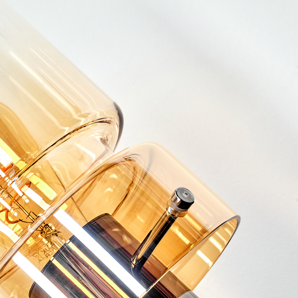 Nahaufnahme einer modernen Lampe mit bernsteinfarbenen Glasschirmen und Metalldetails. Der Hintergrund ist weiß.
