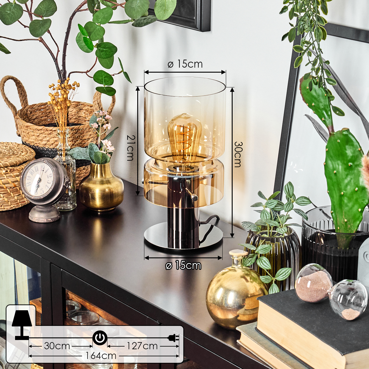Eine dekorative Lampe mit goldener Glühbirne steht auf einem schwarzen Tisch mit verschiedenen Dekorationen und Pflanzen. Abmessungen sind ebenfalls angegeben.