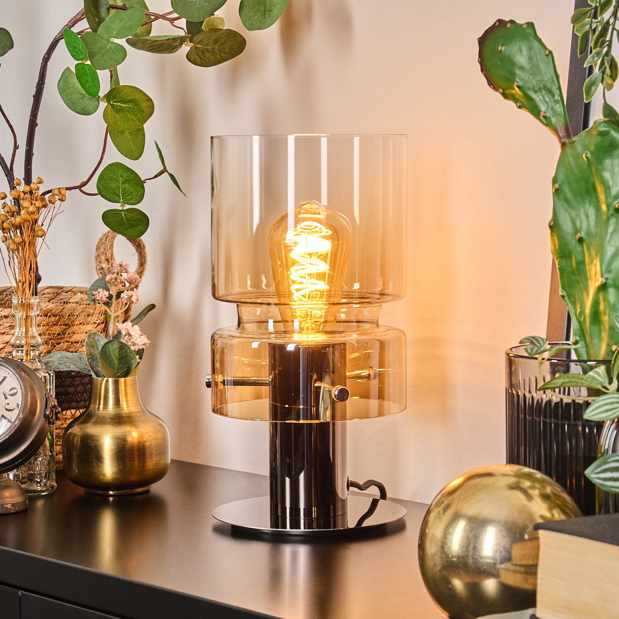 Eine beleuchtete Lampe mit sichtbarer Glühbirne steht auf einem schwarzen Tisch. Dekorative Pflanzen und eine goldene Kugel sind ebenfalls auf dem Tisch.