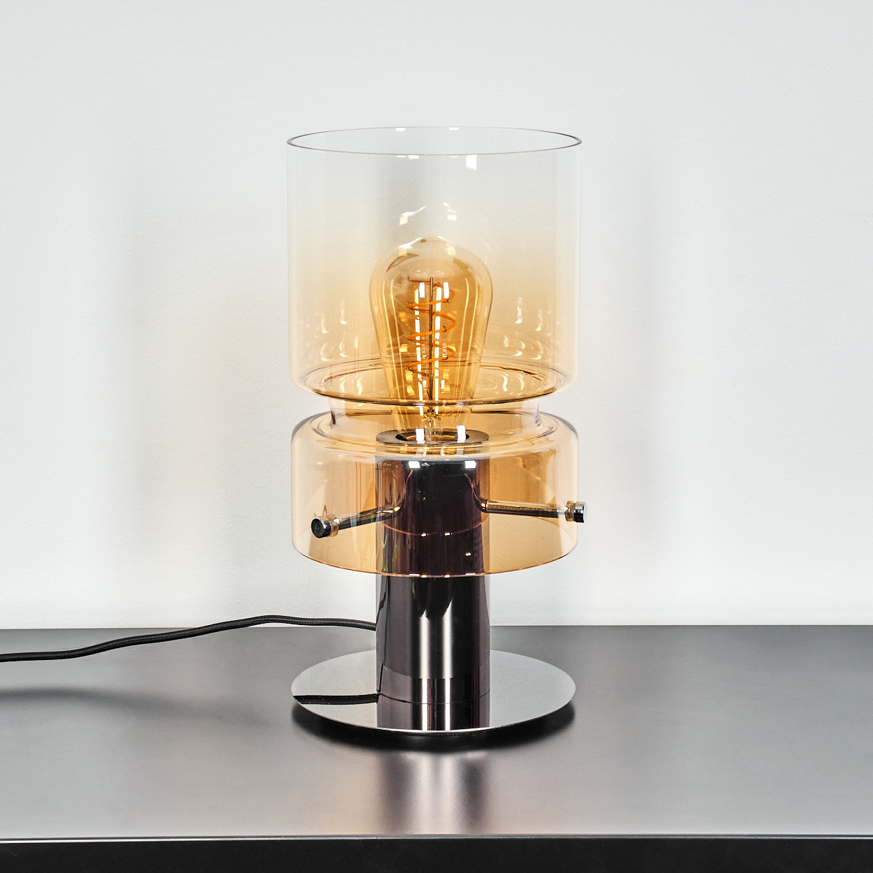 Eine moderne Lampe mit goldener Glühbirne in einem Glasschirm, die auf einer reflektierenden Oberfläche steht. Ein schwarzes Kabel ist sichtbar.