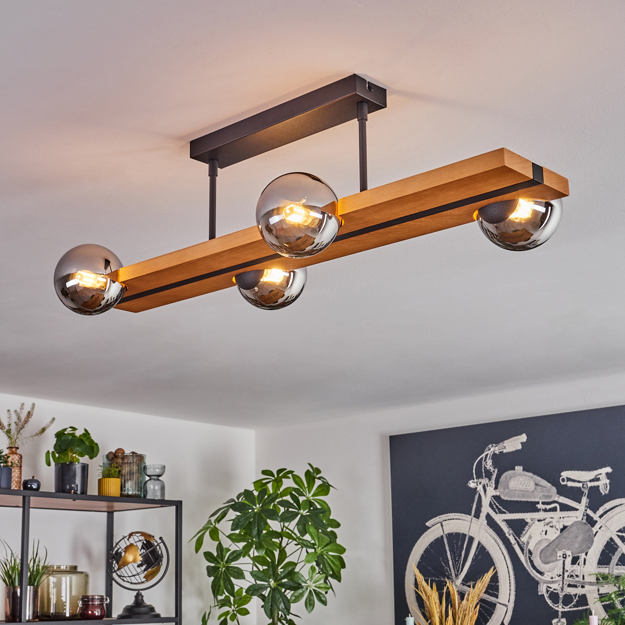 Moderne Deckenlampe aus Holz mit vier Glaskugeln. Pflanzen und ein Fahrradkunstwerk sind im Hintergrund zu sehen.