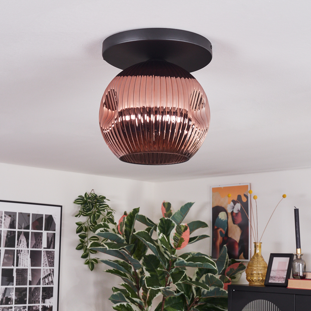 Eine kupferfarbene, kugelförmige Deckenlampe hängt. Darunter sind Pflanzen, Kunst und ein schwarzer Schrank zu sehen.