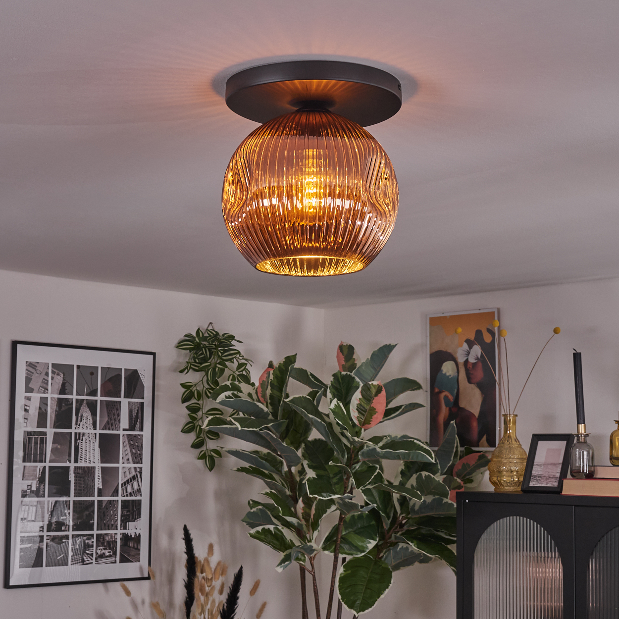 Eine dekorative Deckenlampe mit bernsteinfarbenem Glasschirm hängt in einem Raum. Pflanzen, ein gerahmtes Bild und ein Schrank sind sichtbar.