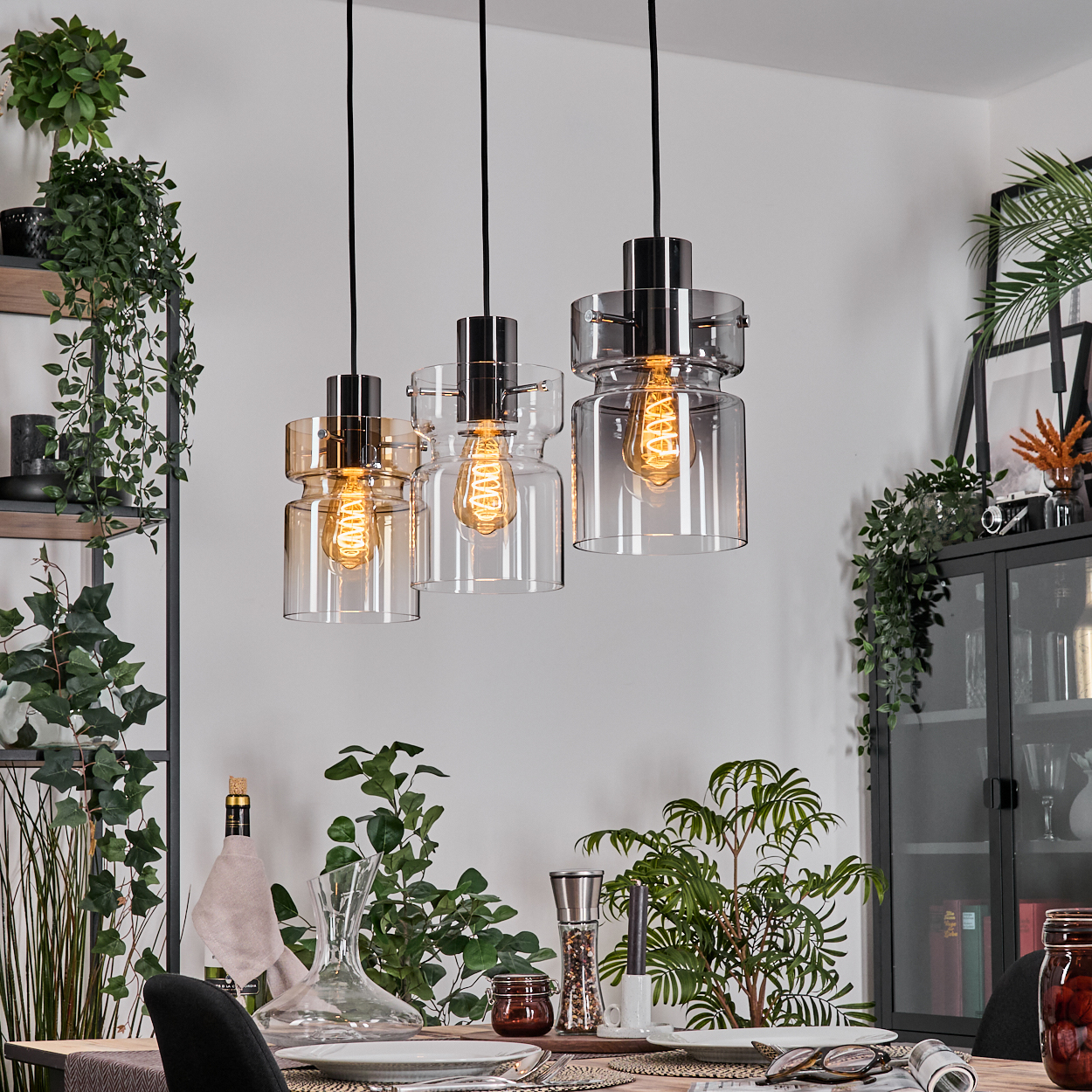 Drei Pendelleuchten mit Edison-Glühbirnen hängen über einem Tisch. Pflanzen und Dekoration umgeben den Tisch und einen Schrank.