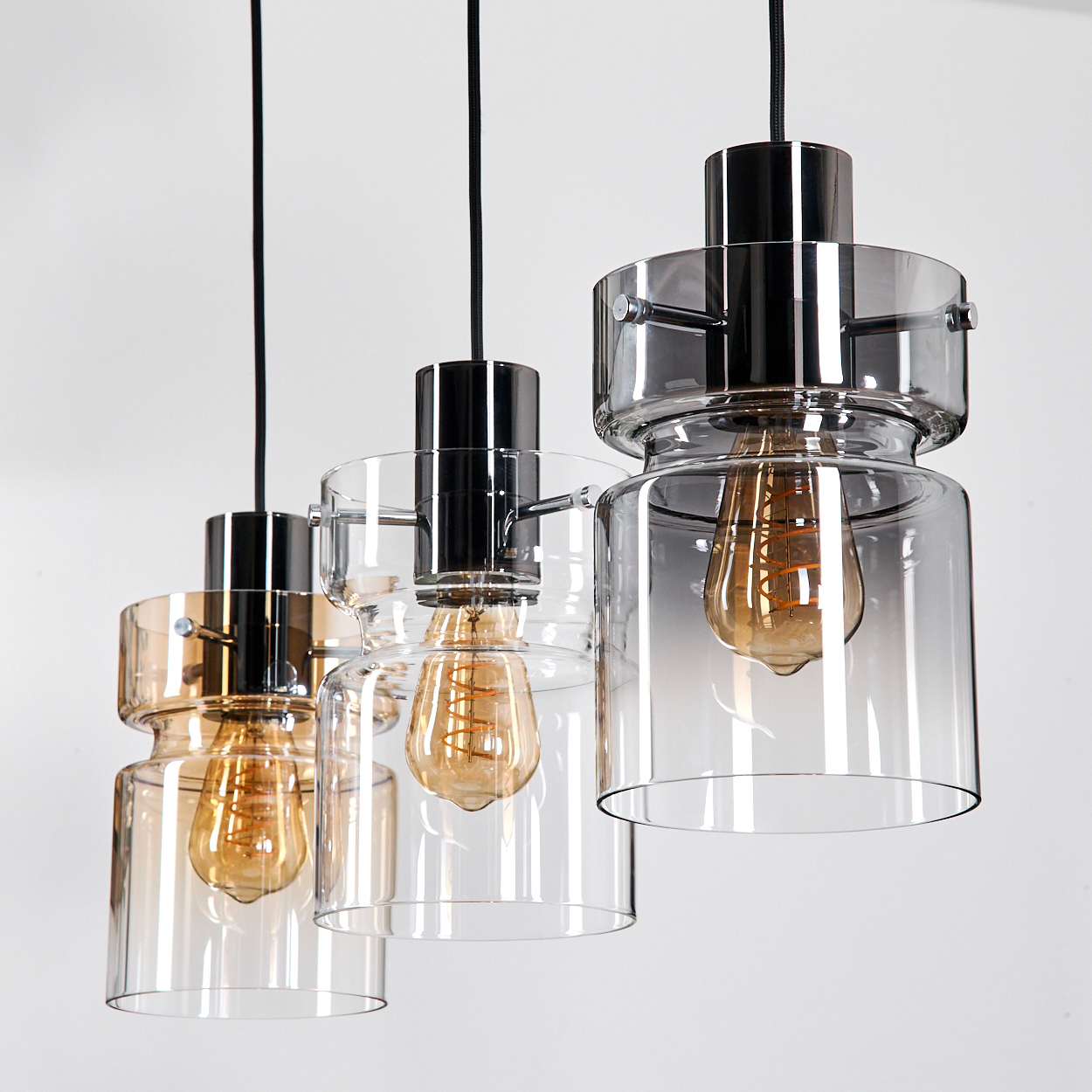 Drei Pendelleuchten mit Glasschirmen und Edison-Glühbirnen hängen vor einem weißen Hintergrund. Jede hat ein schwarzes Kabel und Metallakzente.