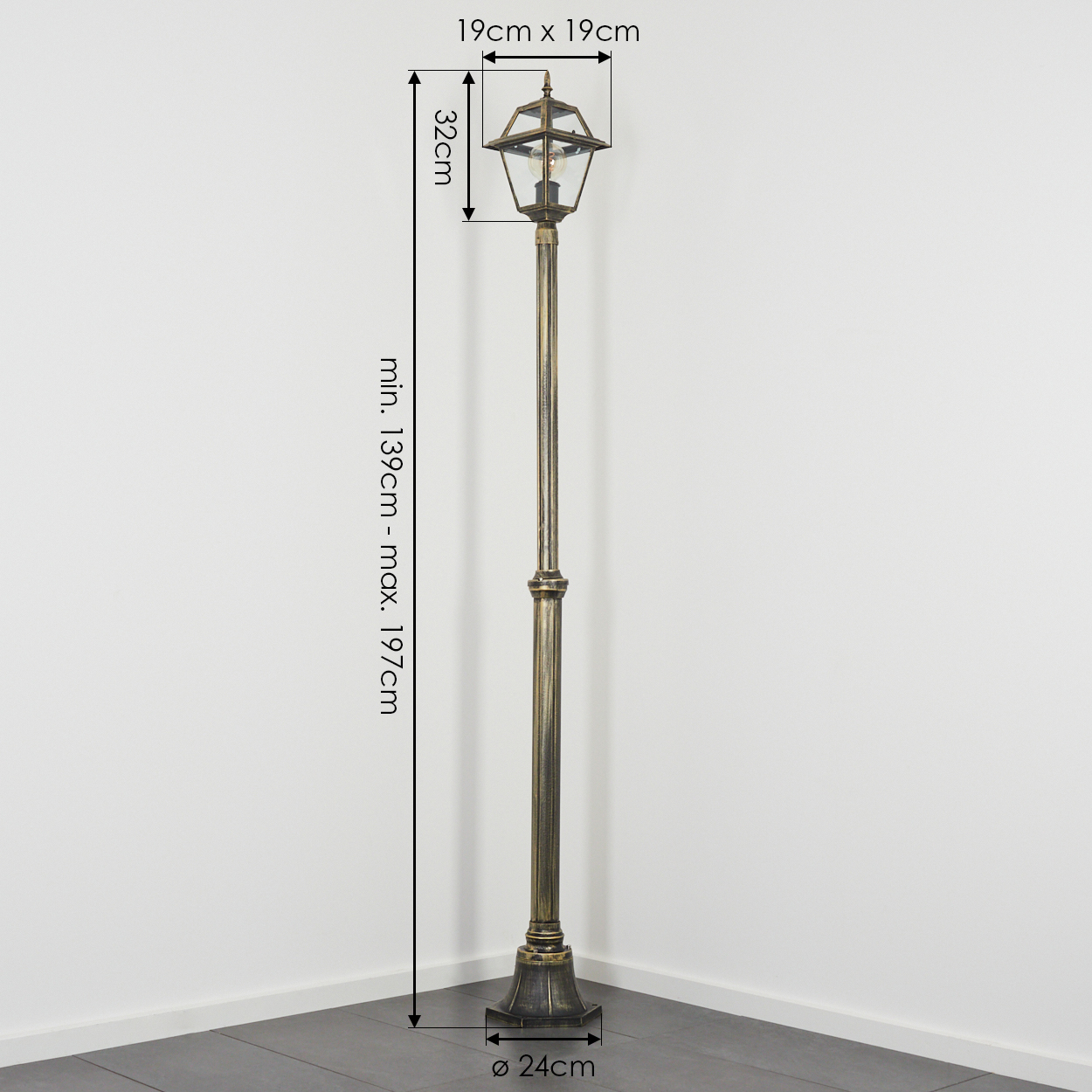Eine bronzefarbene Außenlampe steht vor einer weißen Wand. Abmessungen sind angegeben. Die Basis befindet sich auf einem dunklen Boden. Die Lampe hat ein klassisches Design.