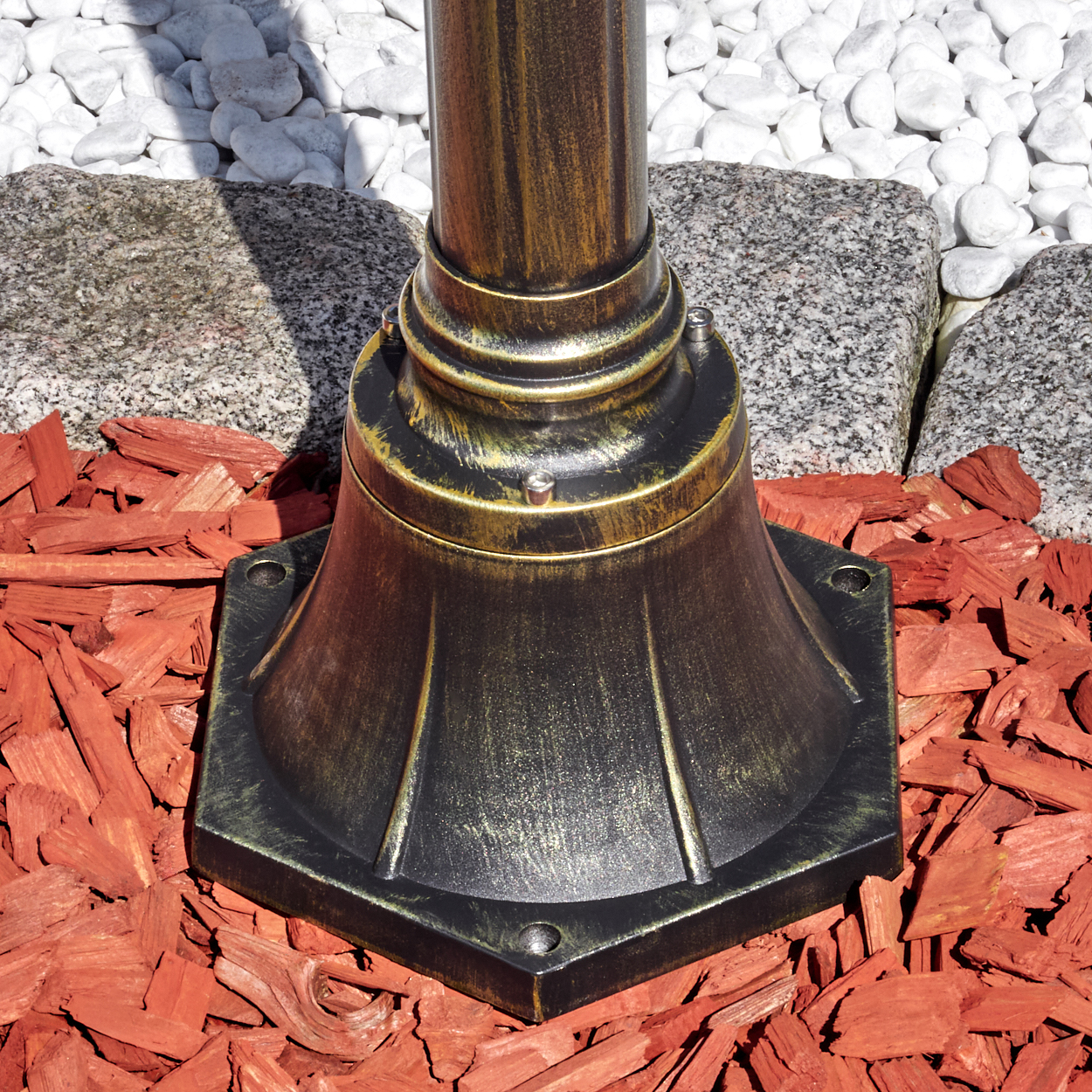Ein bronzefarbener Lampenpfostenfuß steht auf roter Mulch, mit grauen Steinen und weißen Kieselsteinen im Hintergrund.