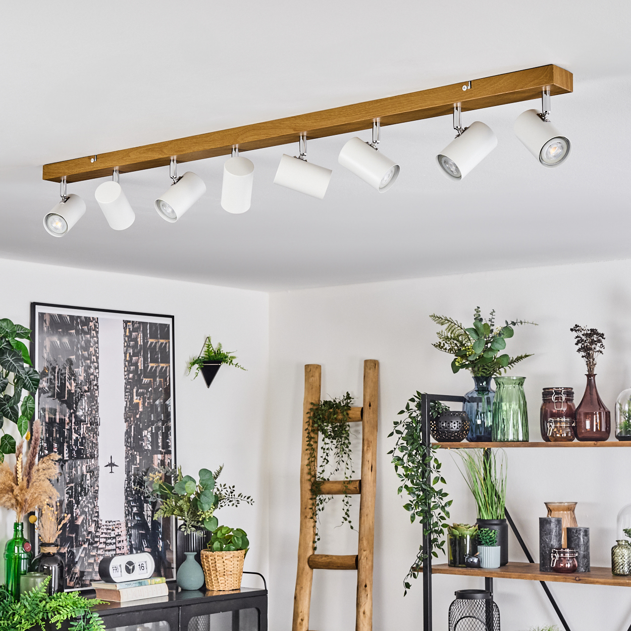 Ein modernes Interieur mit einer hölzernen Deckenleuchte. Darunter sind ein Bild, eine Holzleiter und Regale mit Pflanzen und Vasen zu sehen.