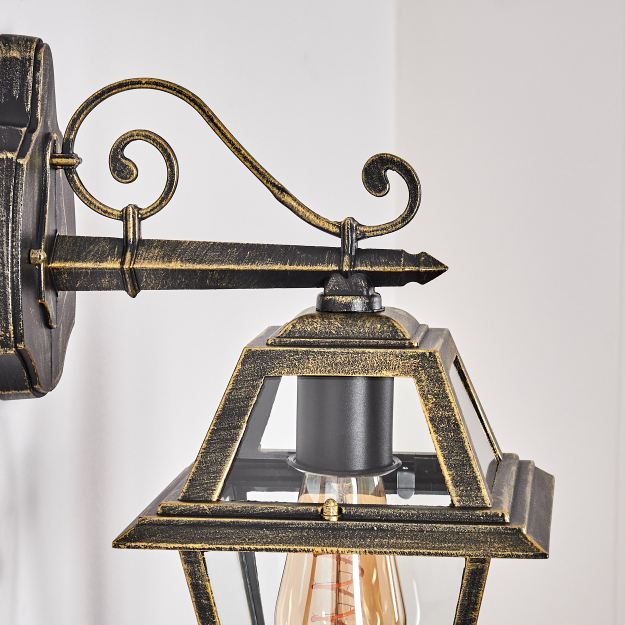 Eine verzierte, bronzefarbene Außenlampe. Sie ist an einer Wand montiert und hat ein glasgeschlossenes Licht mit einer sichtbaren, leuchtenden Glühbirne.
