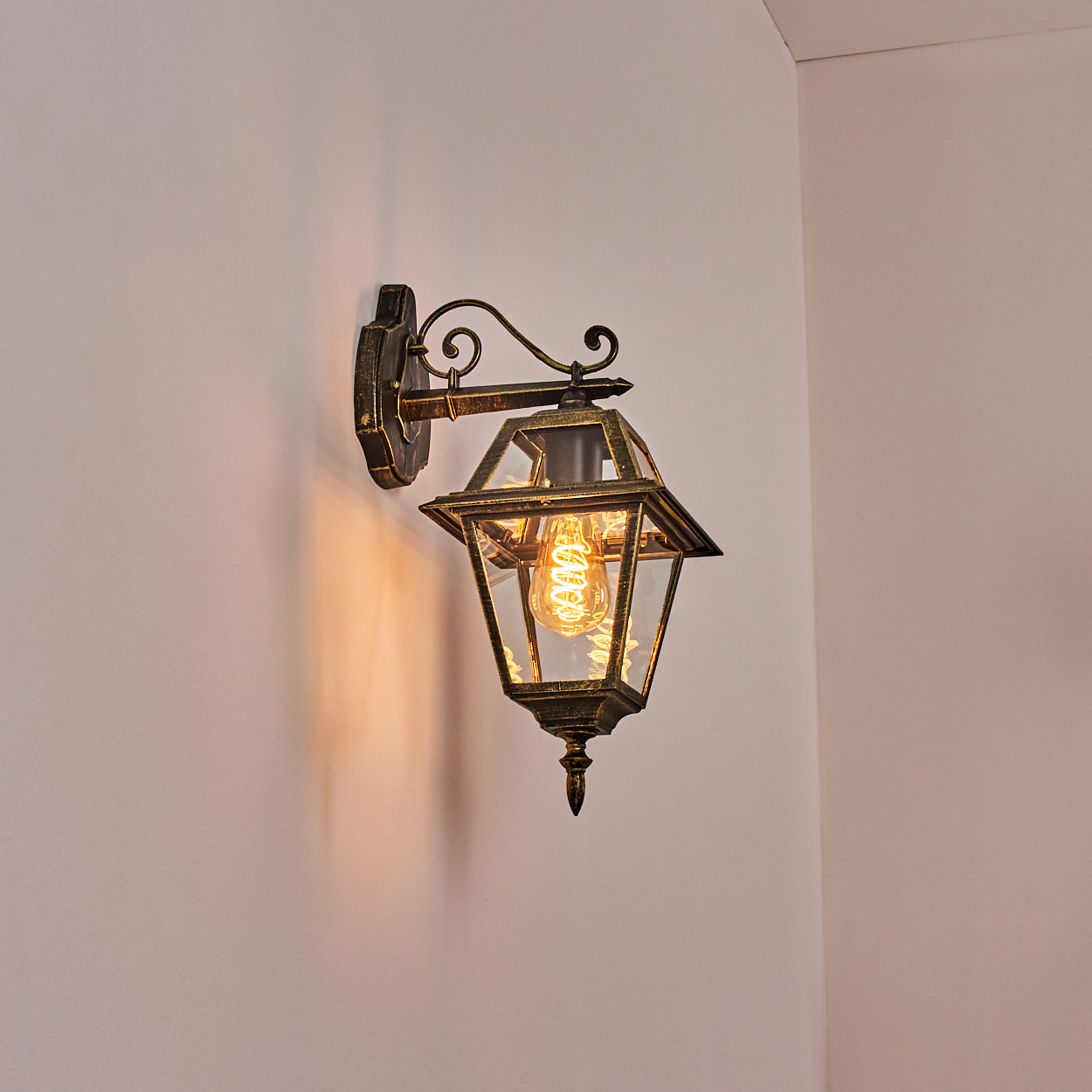 Eine dekorative, an der Wand montierte Laterne mit einer leuchtenden Glühbirne mit Spiralfaden. Sie ist goldfarben vor einer hellrosa Wand.