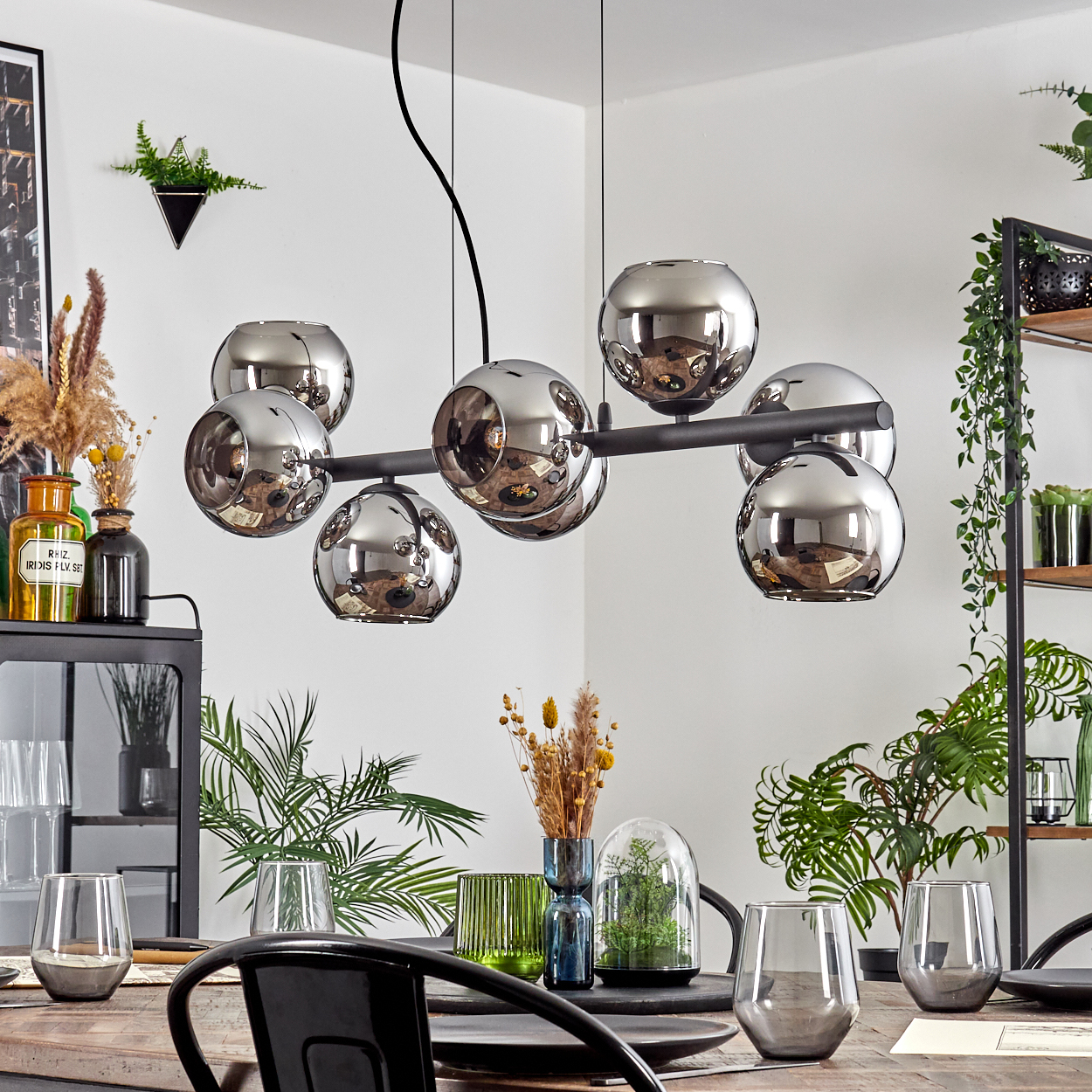 Esszimmer mit Chrom-Leuchte. Pflanzen, Gläser und Tisch im Vordergrund. Regale und Kunst im Hintergrund.
