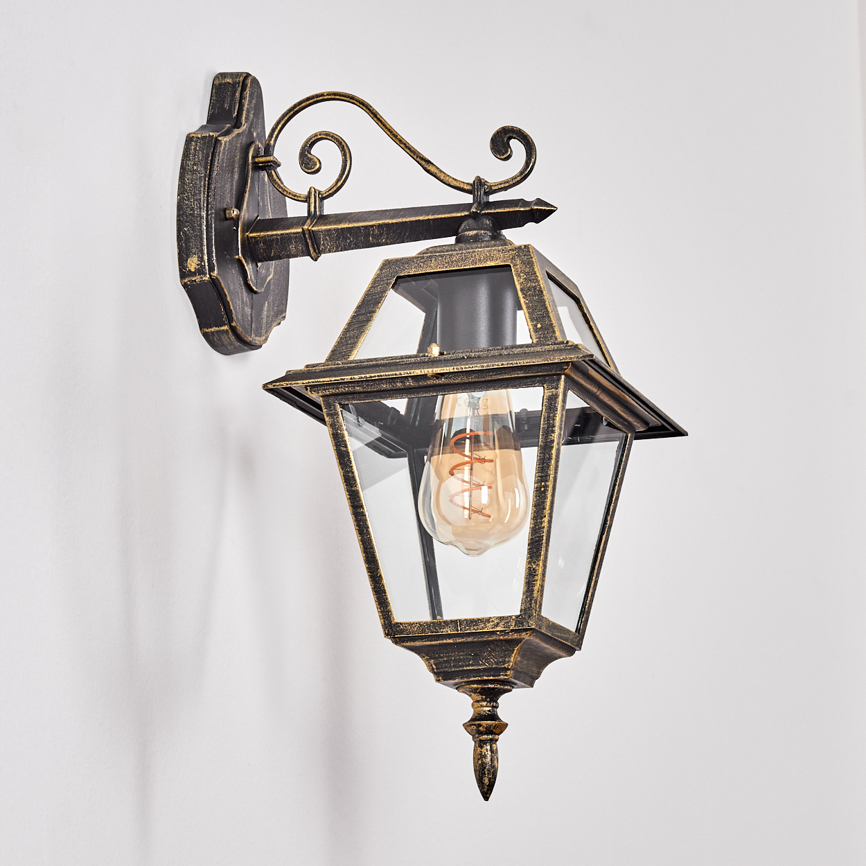Eine verzierte Wandlampe im antiken Stil mit einer Glühbirne in einem Glaskasten. Die Lampe hat eine Gold- und Schwarz-Ausführung, die an einer weißen Wand befestigt ist.