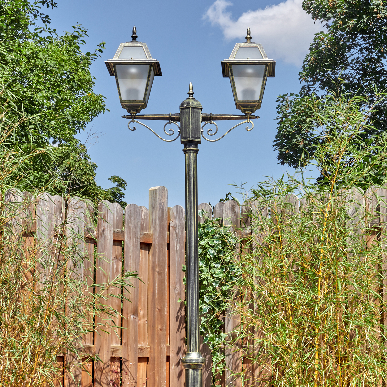 Eine dekorative Laterne mit zwei Lampen steht vor einem Holzzaun. Grüne Pflanzen und Bäume umgeben die Laterne.