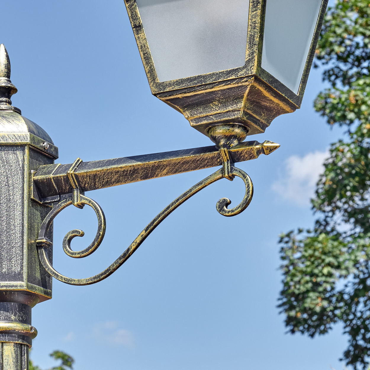Nahaufnahme einer dekorativen Laterne vor blauem Himmel. Die Lampe ist schwarz und gold mit Glasschirm und Baum im Hintergrund.