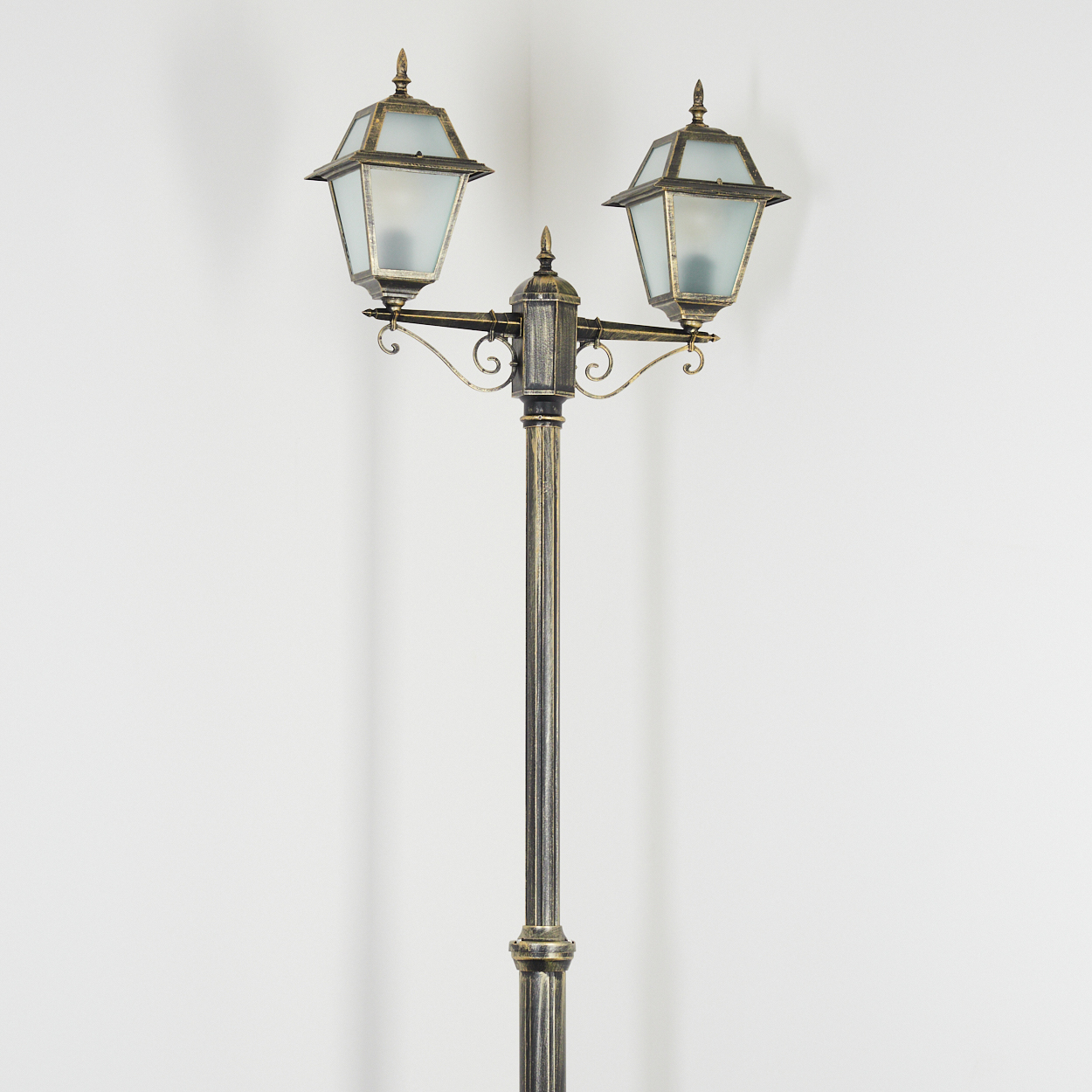 Eine dekorative Straßenlaterne. Sie hat zwei Lampen mit Milchglas, auf einem verzierten, dunklen Metallpfahl, vor einer weißen Wand.
