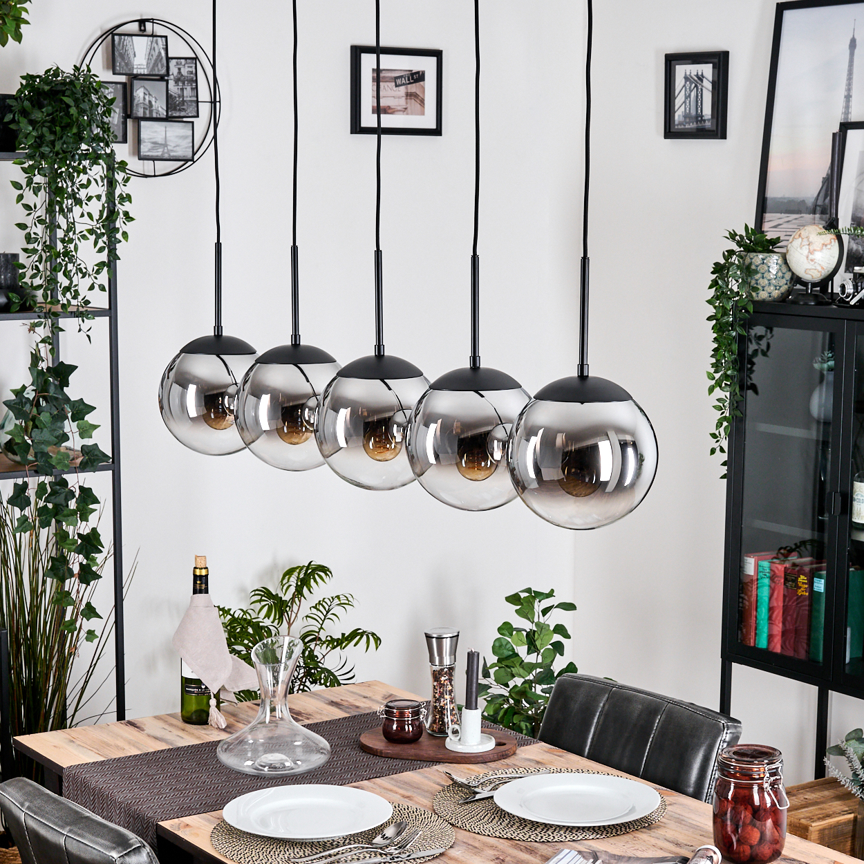 Esszimmer mit Holztisch, Hängelampen und Dekoration. Teller, Wein und Pflanzen stehen auf dem Tisch. Wandkunst und ein schwarzer Schrank sind sichtbar.