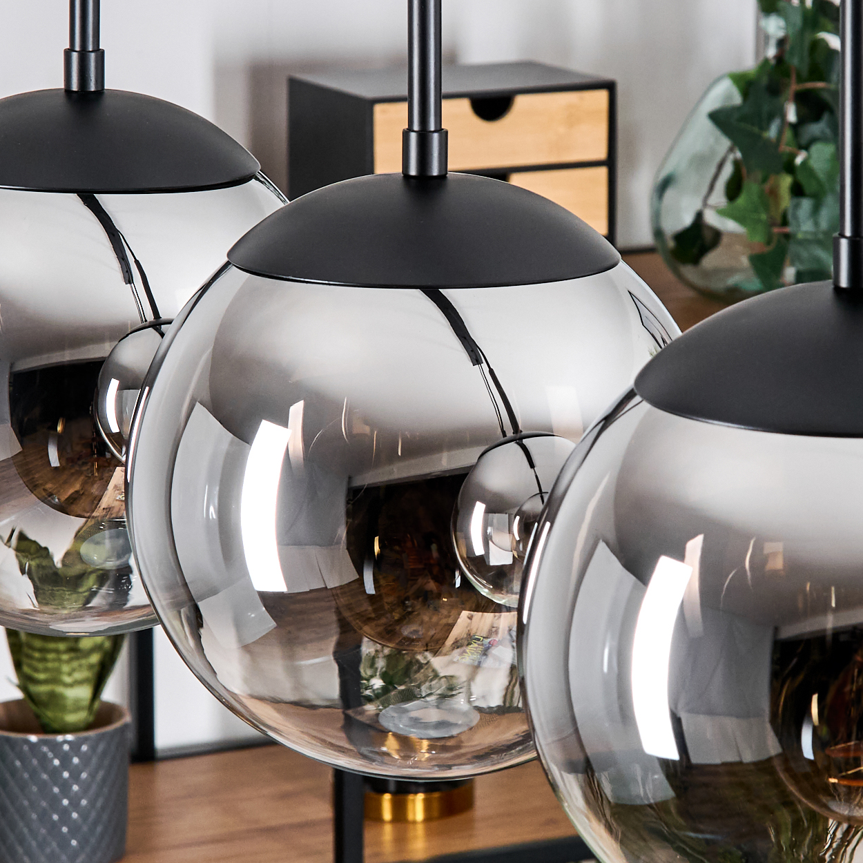 Drei kugelförmige Pendelleuchten mit schwarzen Oberteilen und reflektierendem Glas. Pflanzen und Holzmöbel sind im Hintergrund zu sehen.