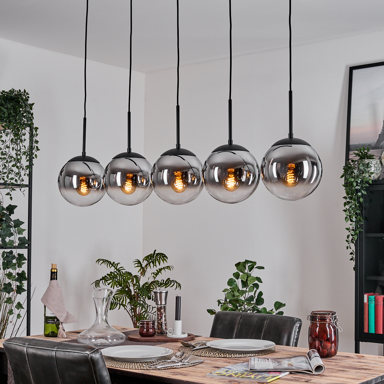 Esszimmer mit einem gedeckten Tisch, über dem fünf Kugelhängeleuchten hängen. Pflanzen und ein gerahmtes Bild befinden sich im Hintergrund.