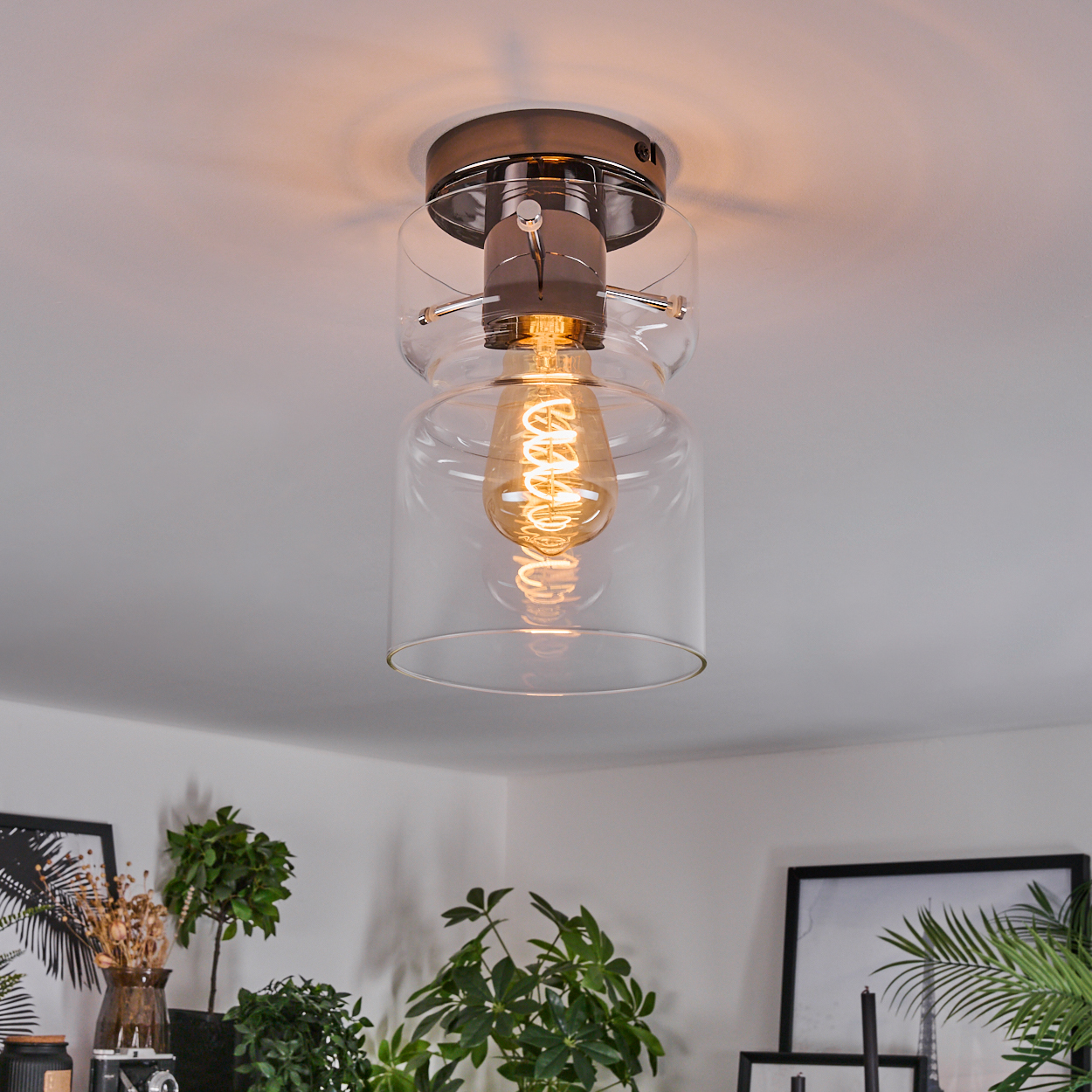 Eine moderne Deckenleuchte mit sichtbarer Glühbirne in einem klaren Glasschirm, umgeben von einer weißen Decke und Pflanzen darunter.