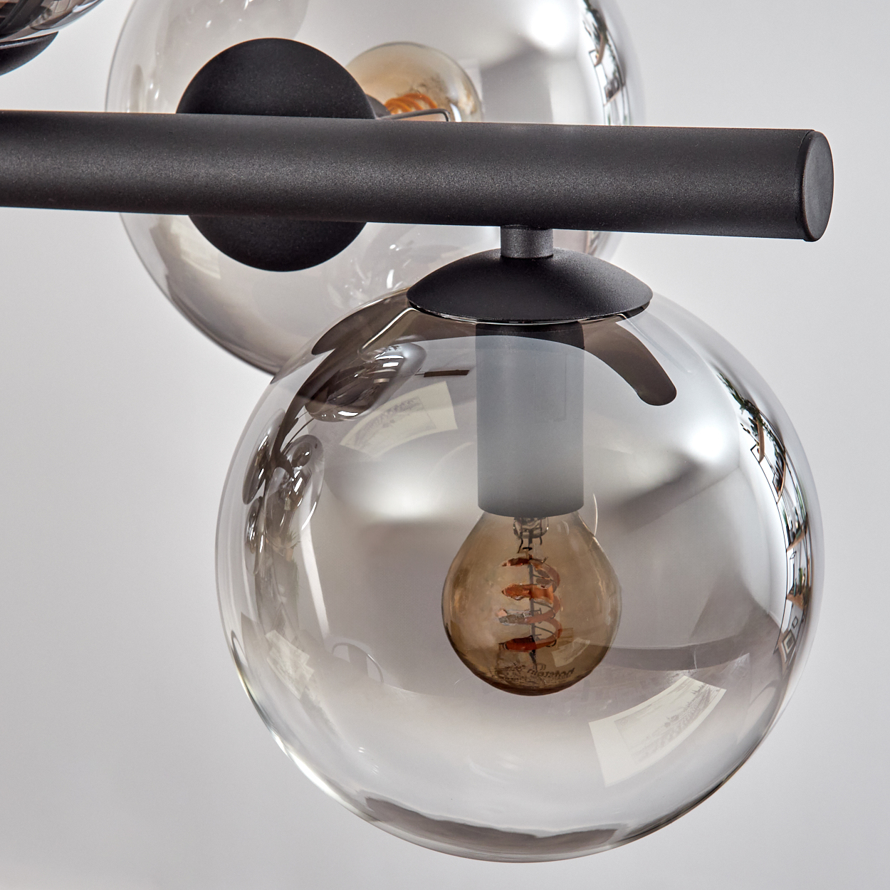 Nahaufnahme einer modernen Leuchte mit drei klaren, kugelförmigen Glasschirmen. Jedes enthält eine warm getönte Glühbirne. Schwarzer Metallrahmen.