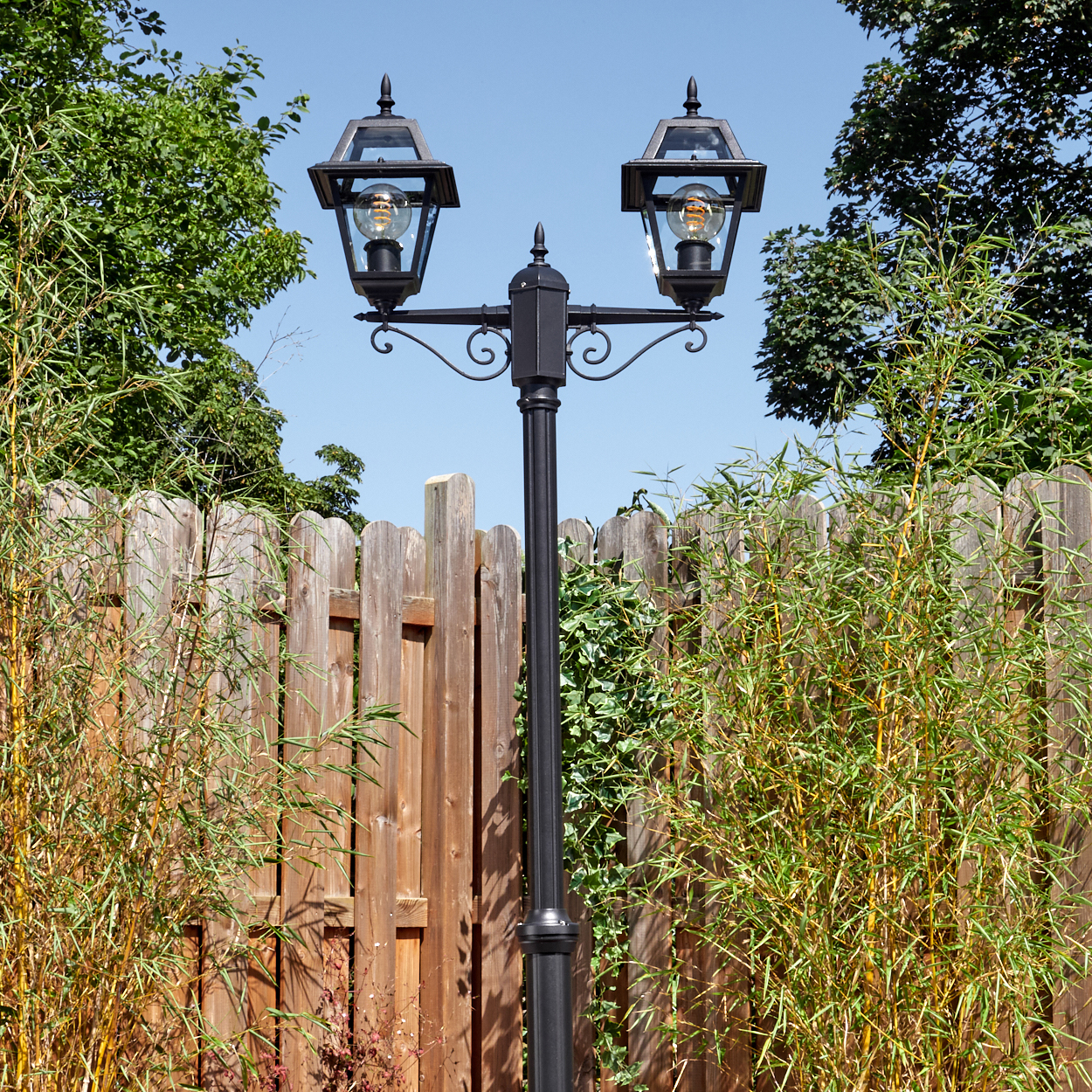 Eine schwarze Laterne mit zwei Lampen steht vor einem Holzzaun. Bambus und Bäume flankieren den Zaun unter blauem Himmel.