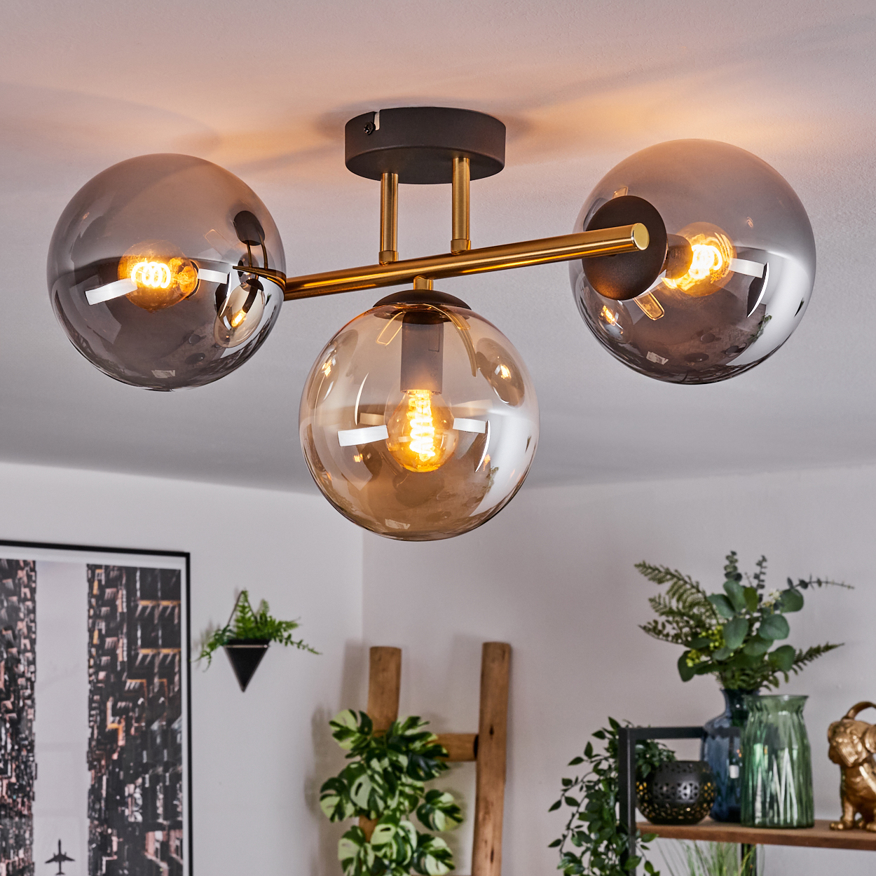 Eine moderne Deckenlampe mit drei kugelförmigen Schirmen. Die Schirme sind aus Glas mit sichtbaren Glühbirnen. Pflanzen und Dekorationsgegenstände sind im Hintergrund.