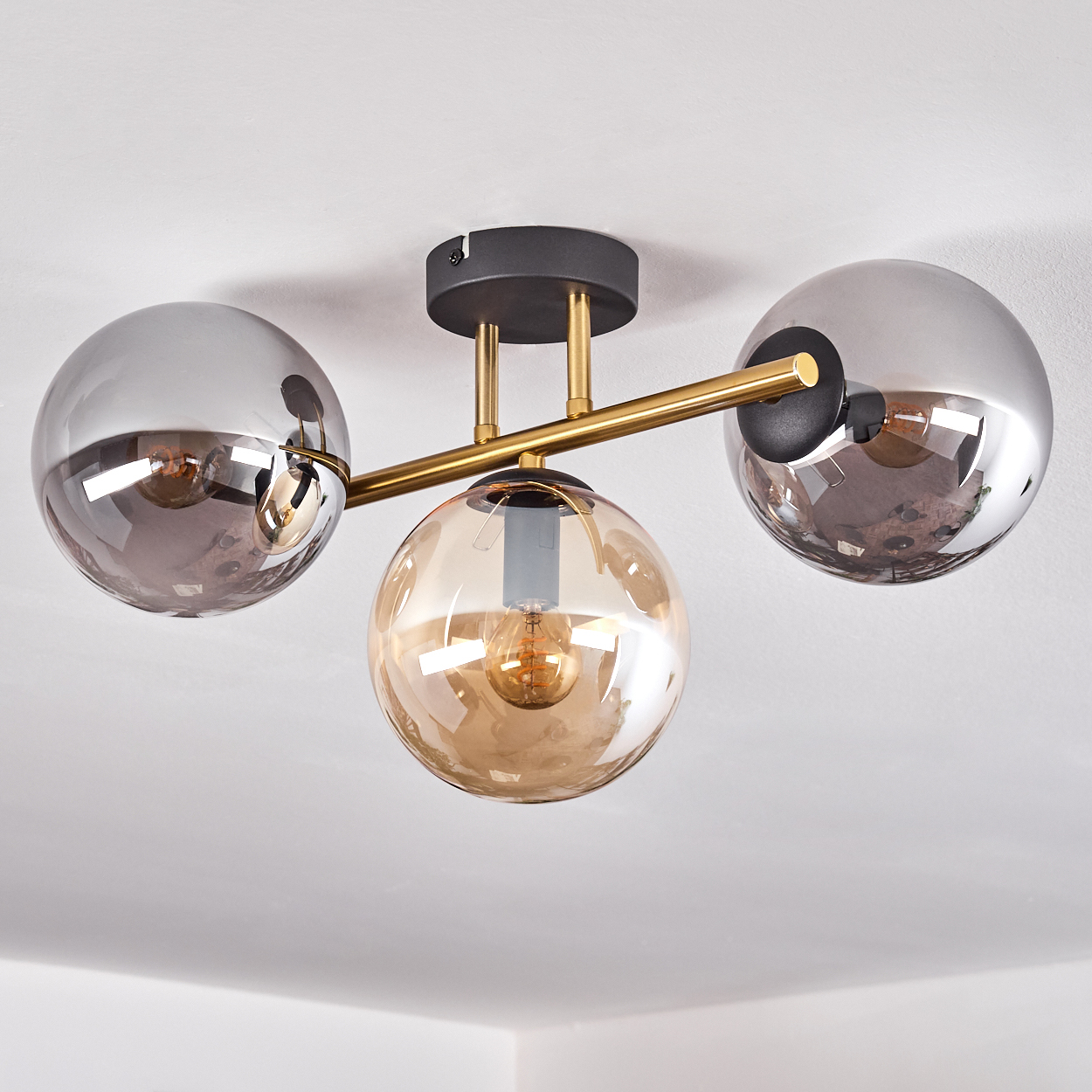 Eine moderne Deckenleuchte mit drei Glaskugeln in Silber und Bernstein, montiert an einem goldfarbenen Rahmen, vor weißer Decke.