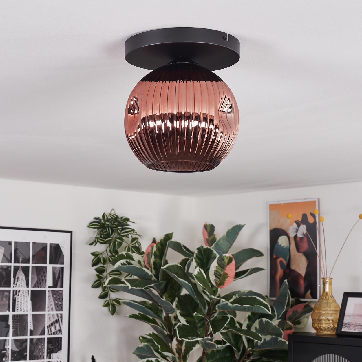 Eine kupferfarbene, geriffelte Glasdeckenlampe hängt an einer weißen Decke. Pflanzen und Kunstwerke sind unter der Lampe zu sehen.