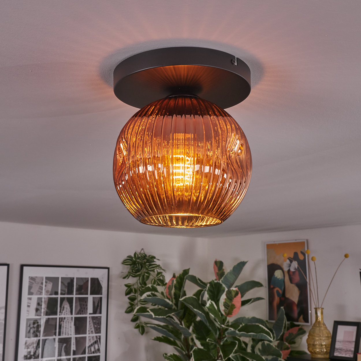 Eine Deckenleuchte mit einer bernsteinfarbenen Glaskugel. Das Licht ist an und beleuchtet den Raum. Pflanzen und Kunstwerke sind unten zu sehen.
