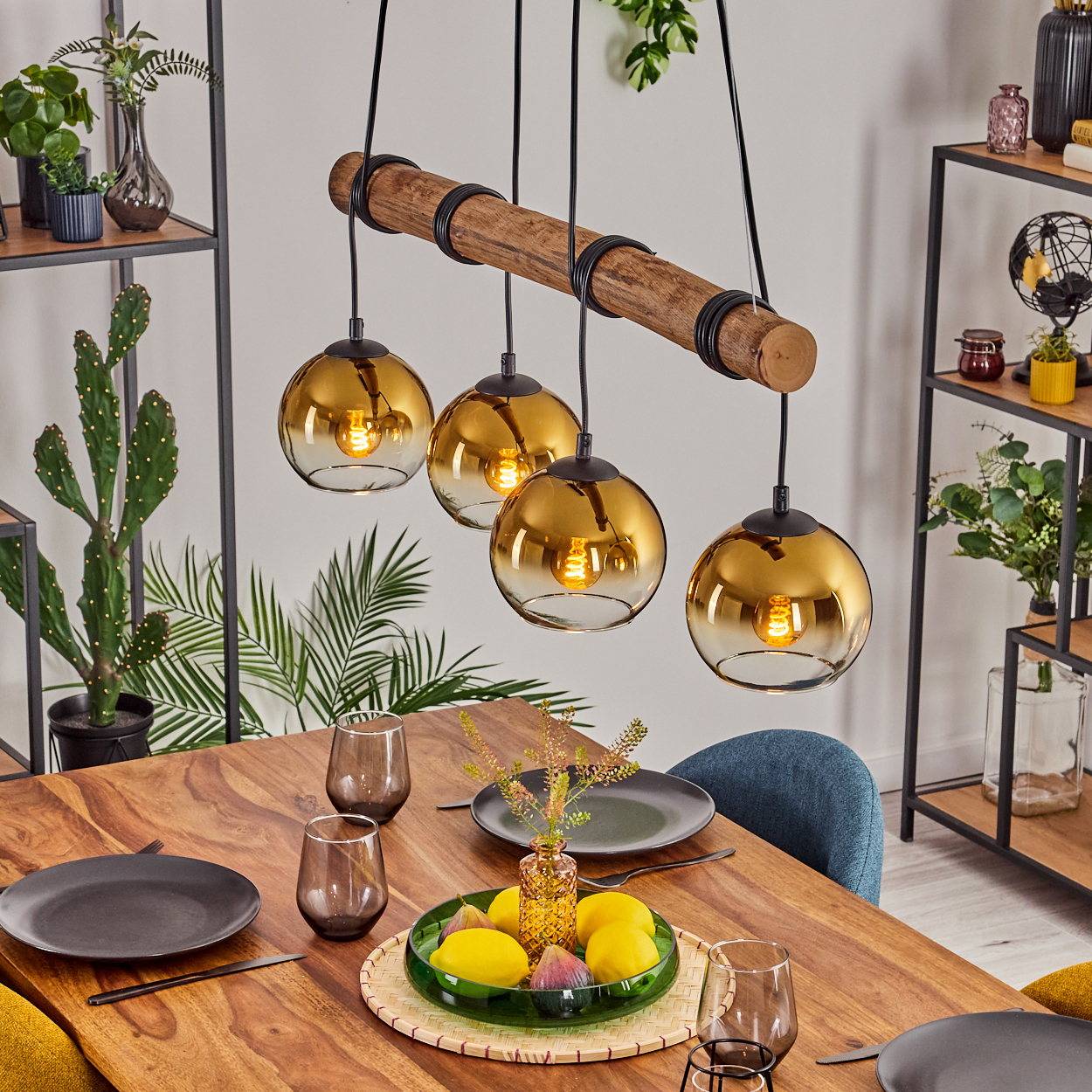 Ein Esstisch mit Holzplatte, dekoriert mit Tellern, Gläsern und einem Mittelstück aus Zitronen und Feigen, beleuchtet von einer Pendelleuchte.