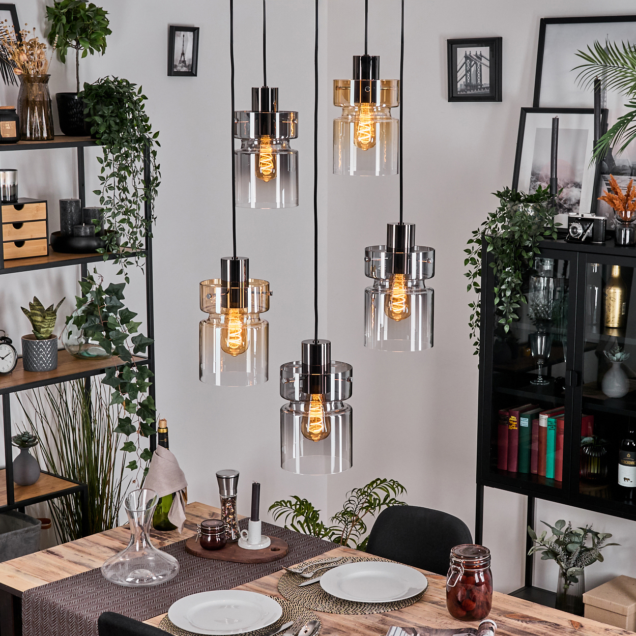 Esszimmer mit Hängelampen. Tisch mit Tellern, Gläsern und Dekorationen. Regale mit Pflanzen und Dekor im Hintergrund.