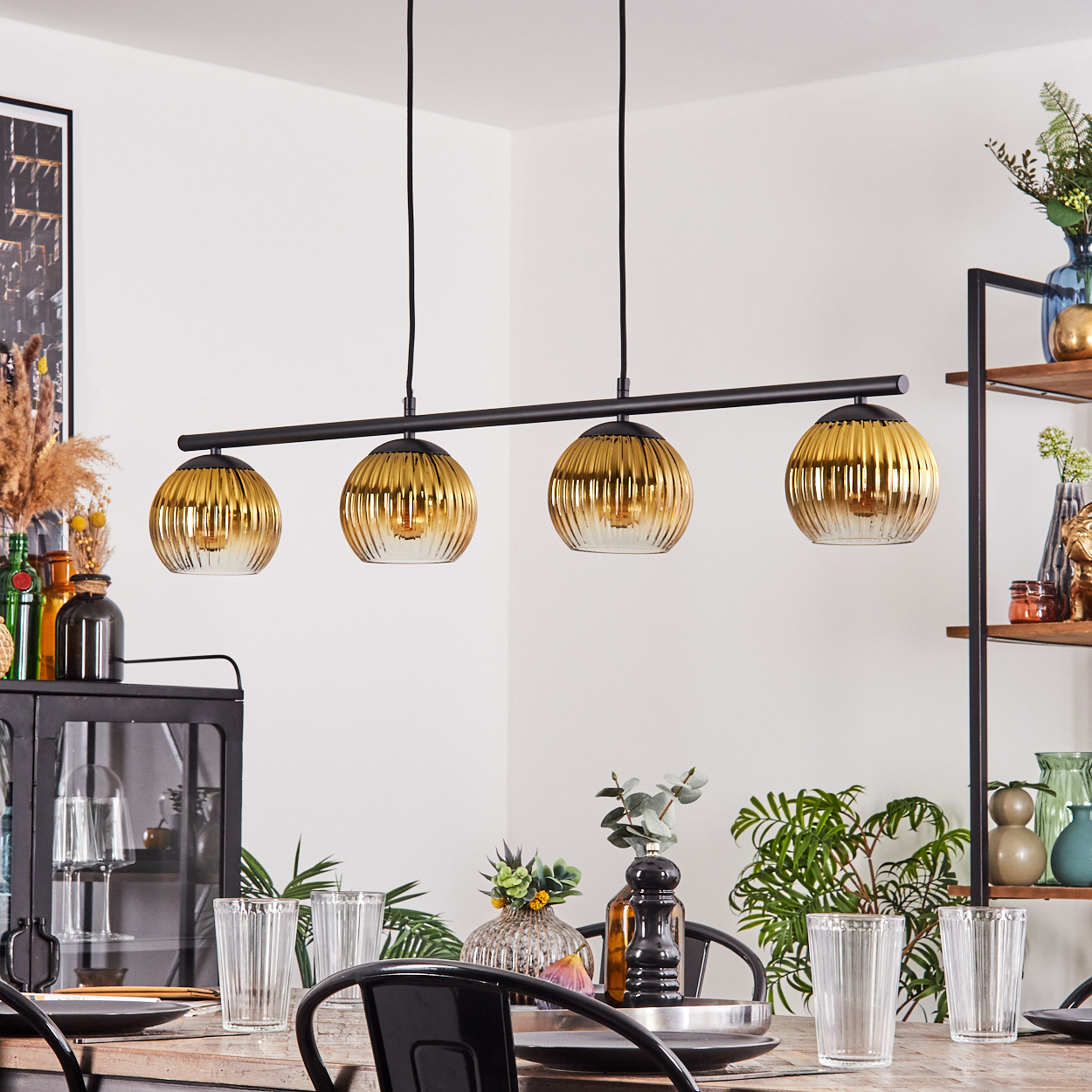 Esszimmer mit gedecktem Tisch. Über dem Tisch hängen vier goldene Glas-Pendelleuchten. Auf Regalen gibt es Dekorationen.