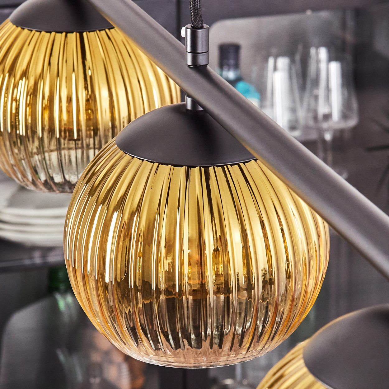 Nahaufnahme von drei goldenen, gerippten Glas-Pendelleuchten vor einem verschwommenen Hintergrund mit Küchenutensilien, in einem Haus.