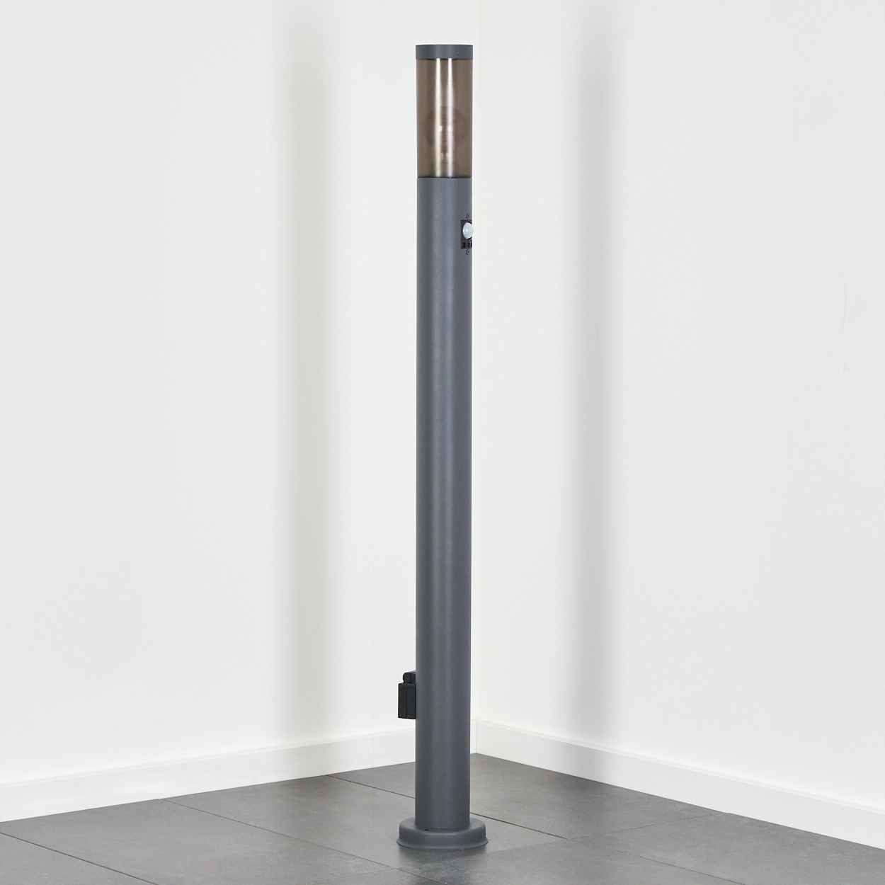 Graue zylindrische Poller mit metallischem Oberteil stehen in einer Ecke vor weißen Wänden und einem grauen Fliesenboden.