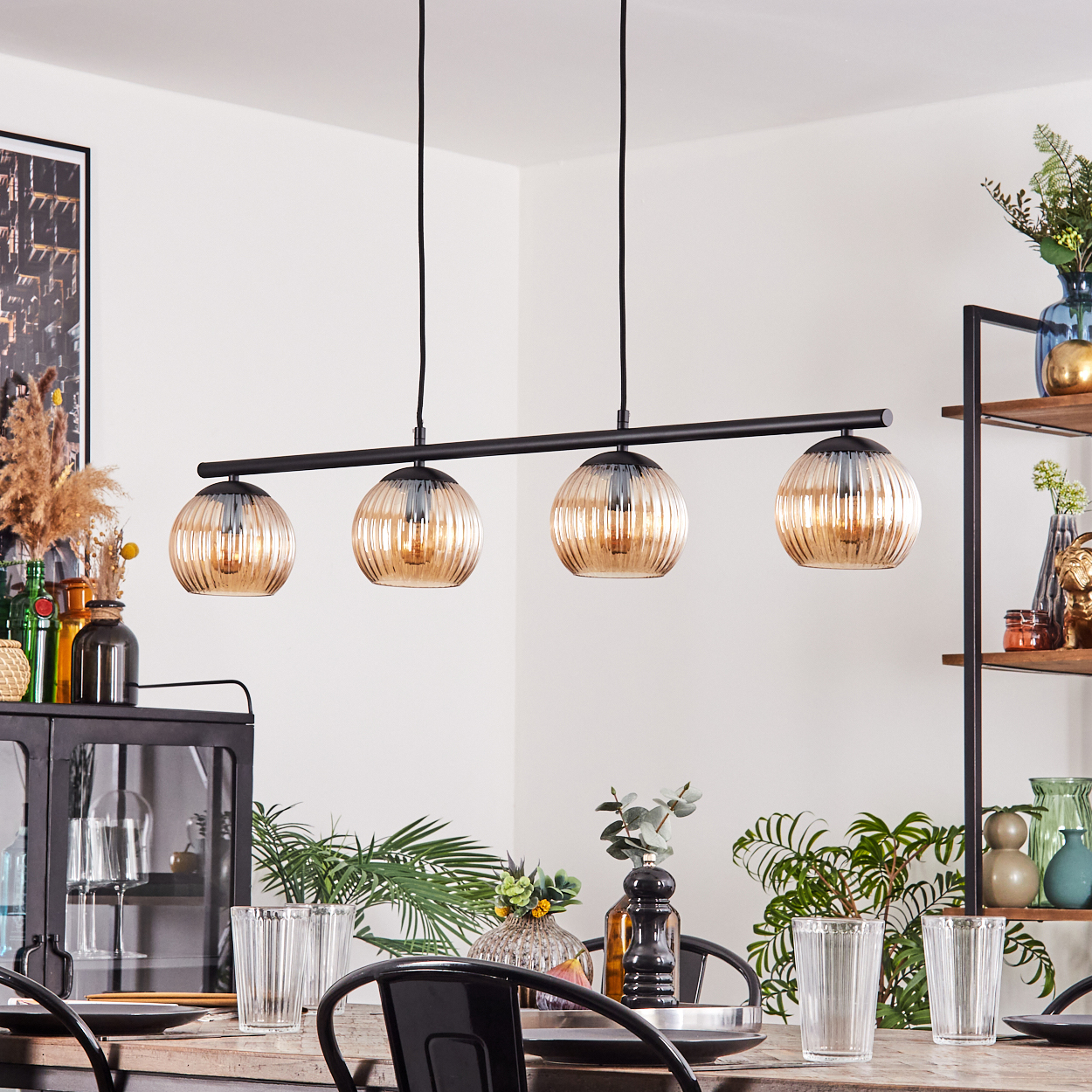 Esszimmer mit gedecktem Tisch. Vier Lampen hängen darüber. Regale und Pflanzen sind im Hintergrund sichtbar.