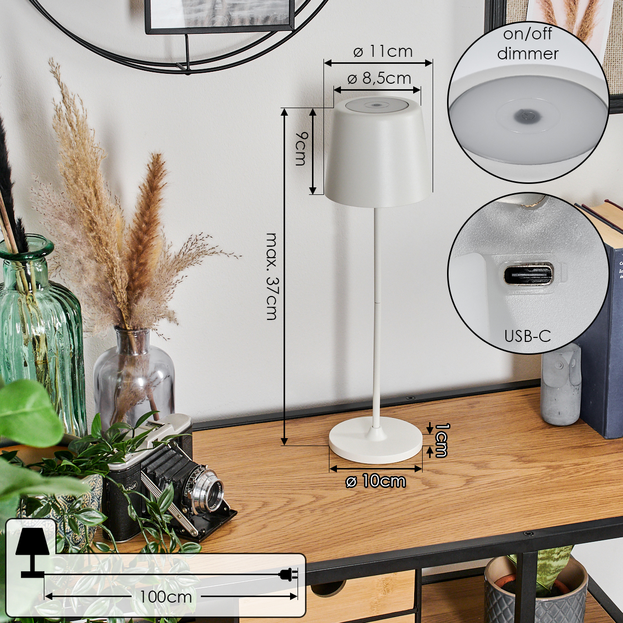 Eine weiße Schreibtischlampe mit zylindrischem Lampenschirm, auf einem Holztisch mit Dekorationsartikeln und Pflanzen.