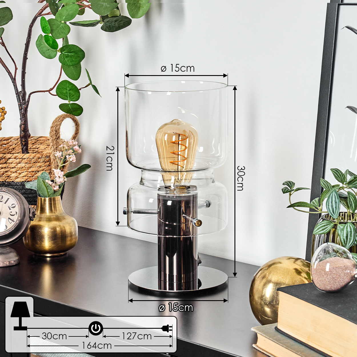 Eine moderne Lampe mit sichtbarer Glühbirne steht auf einer schwarzen Oberfläche. Weitere Objekte sind eine Uhr, eine goldene Vase, Pflanzen und Bücher.