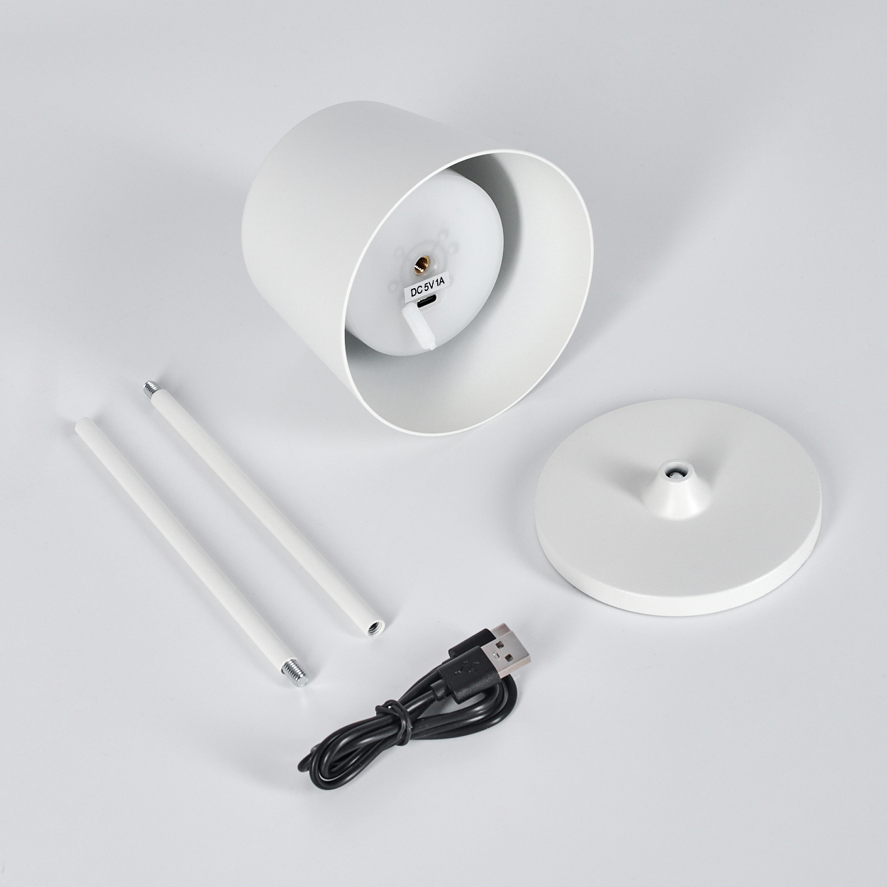 Eine zerlegte weiße Lampe mit Sockel, zwei Stäben, einem zylindrischen Schirm und einem USB-Kabel, alles auf einer weißen Oberfläche.