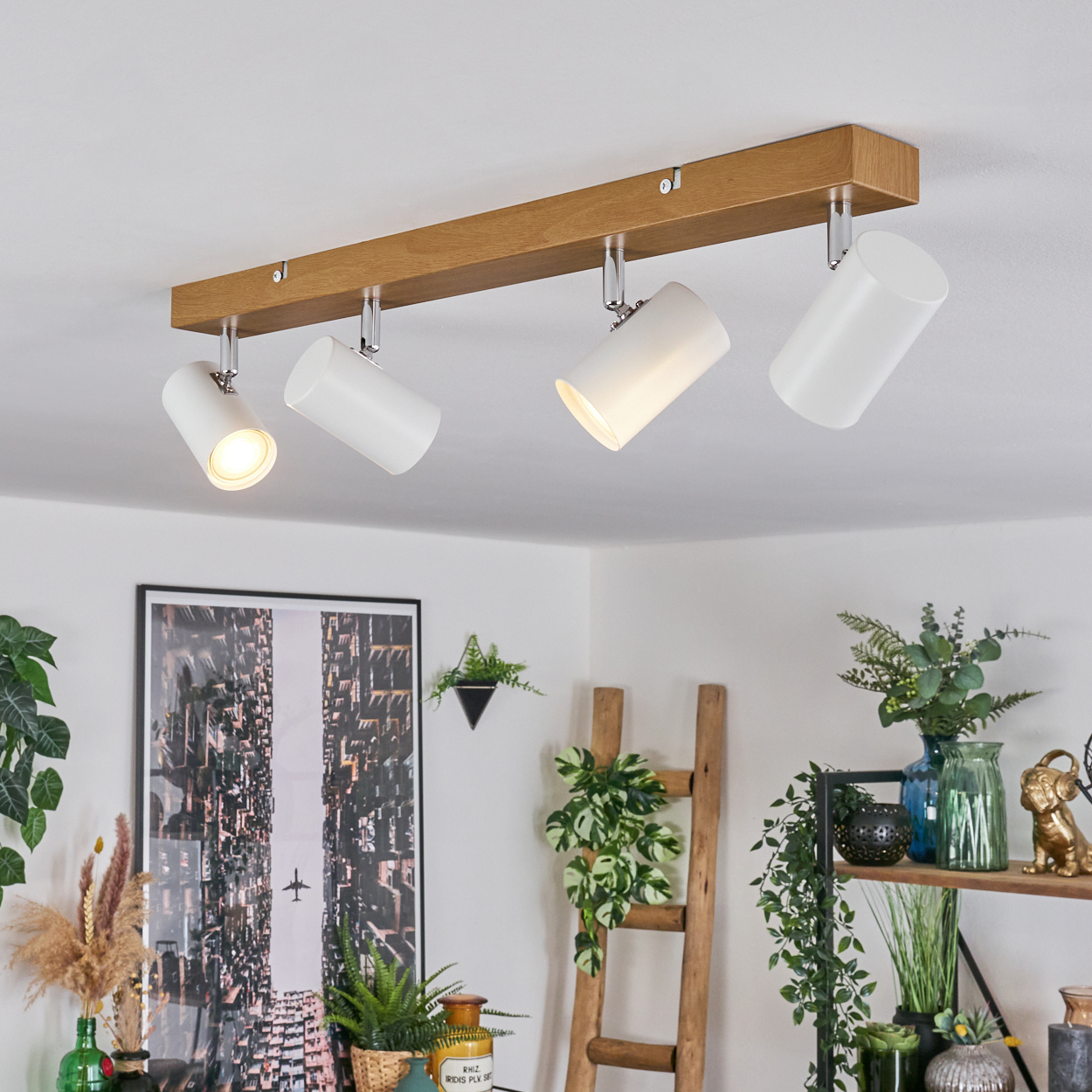 Eine moderne Deckenleuchte mit vier weißen Strahlern auf einem Holzbalken. Pflanzen und Dekoration sind im Hintergrund. Der Raum ist gut beleuchtet.
