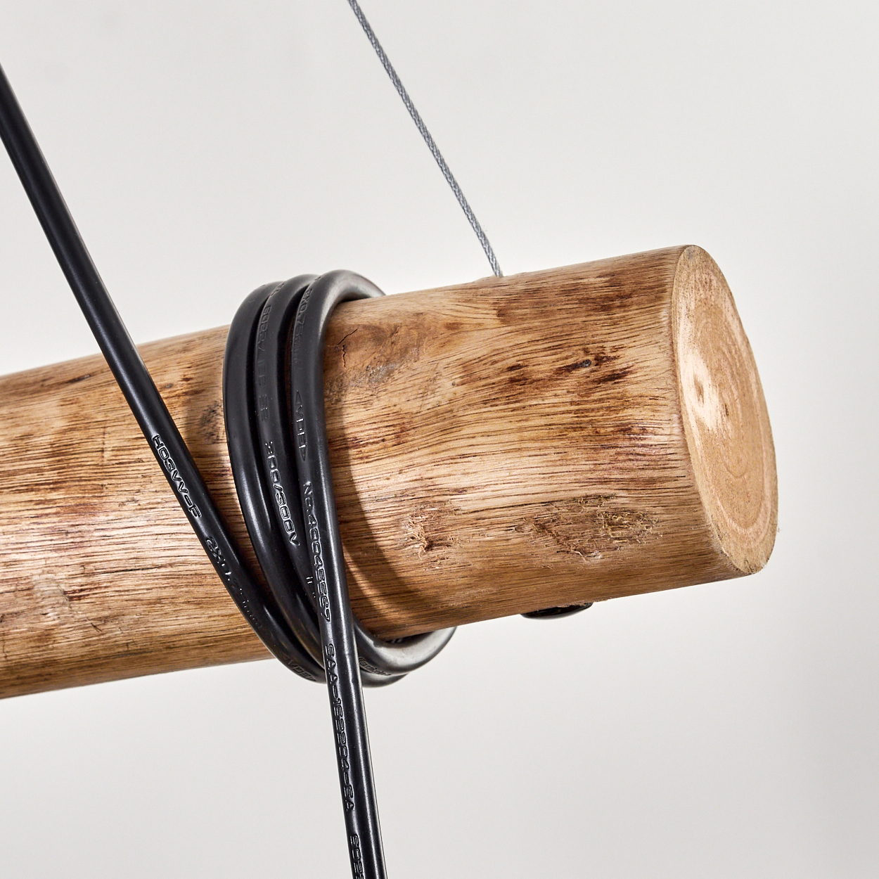 Ein Holzstamm mit einem schwarzen Kabel, das darum gewickelt ist, an einem Draht vor weißem Hintergrund aufgehängt. Das Ende des Stammes ist sichtbar.