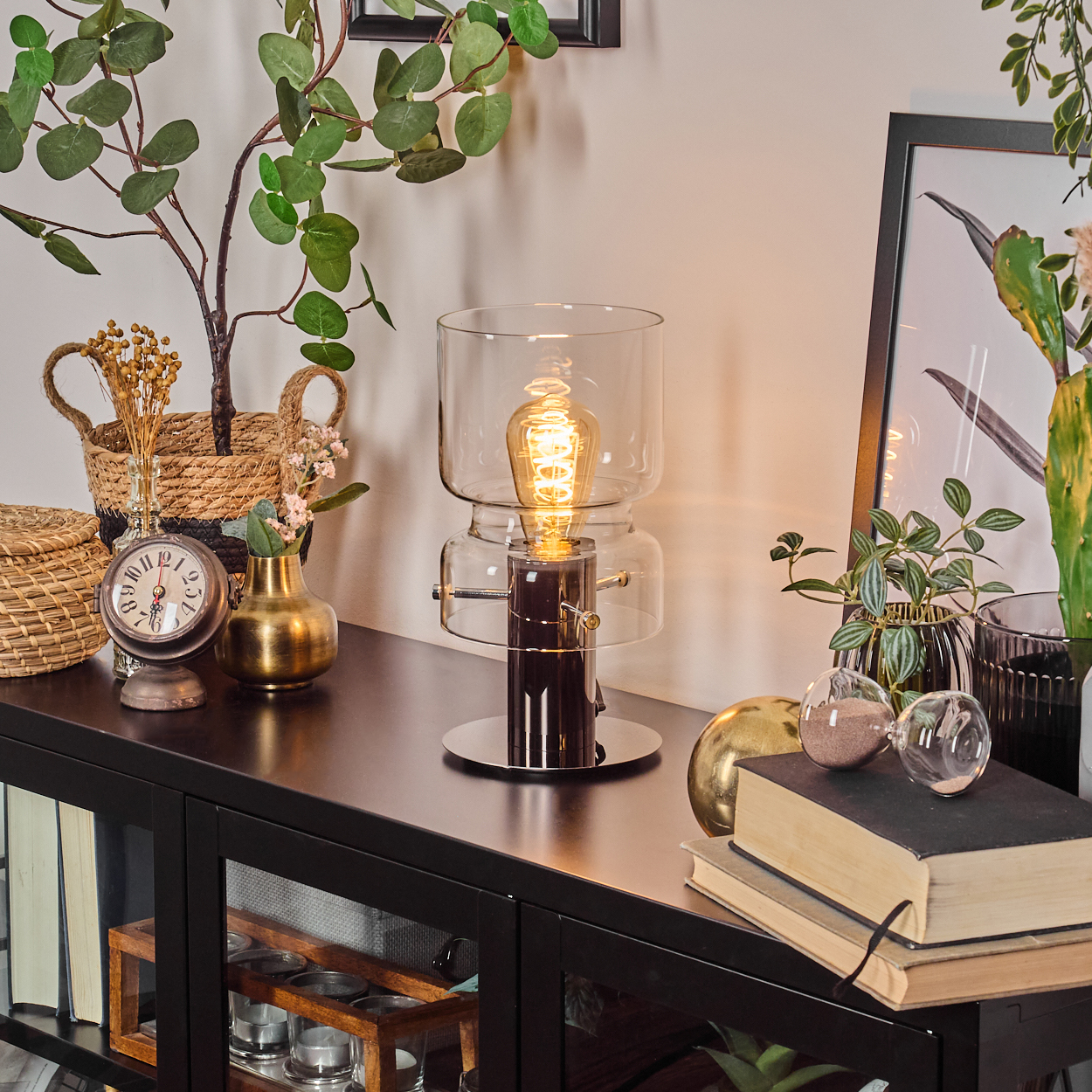 Eine beleuchtete Lampe steht auf einem schwarzen Tisch mit Büchern, Pflanzen und Dekoration. Eine Uhr, eine Vase und ein Bilderrahmen sind ebenfalls sichtbar.