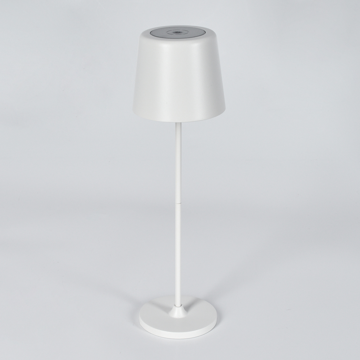 Eine weiße Lampe mit konischem Schirm, dünnem Stiel und rundem Fuß steht auf weißem Untergrund. Der Hintergrund ist weiß.