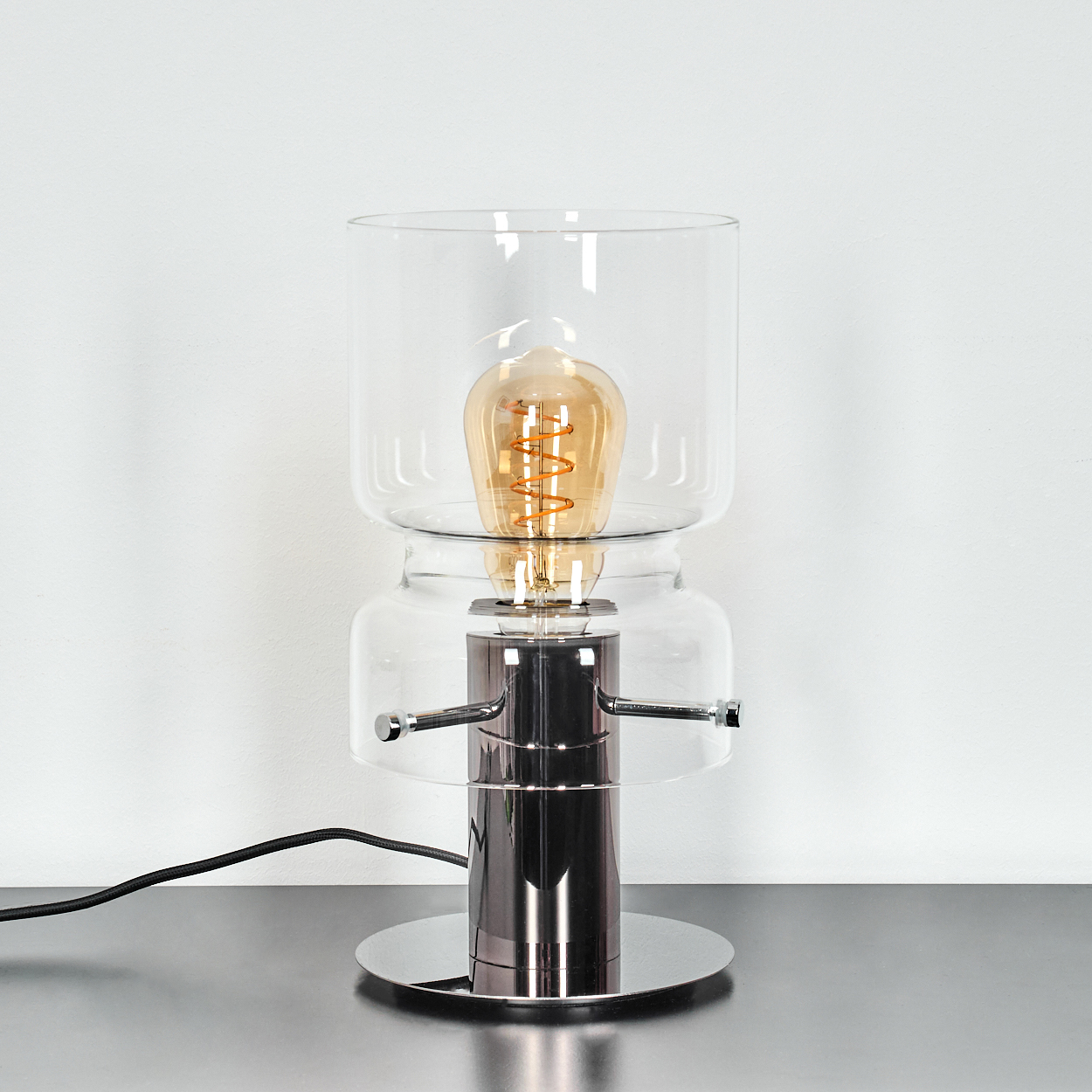 Eine moderne Lampe mit Chromfuß, Glasschirmen und einer sichtbaren, warm getönten Glühbirne. Ein schwarzes Kabel geht vom Fuß aus.