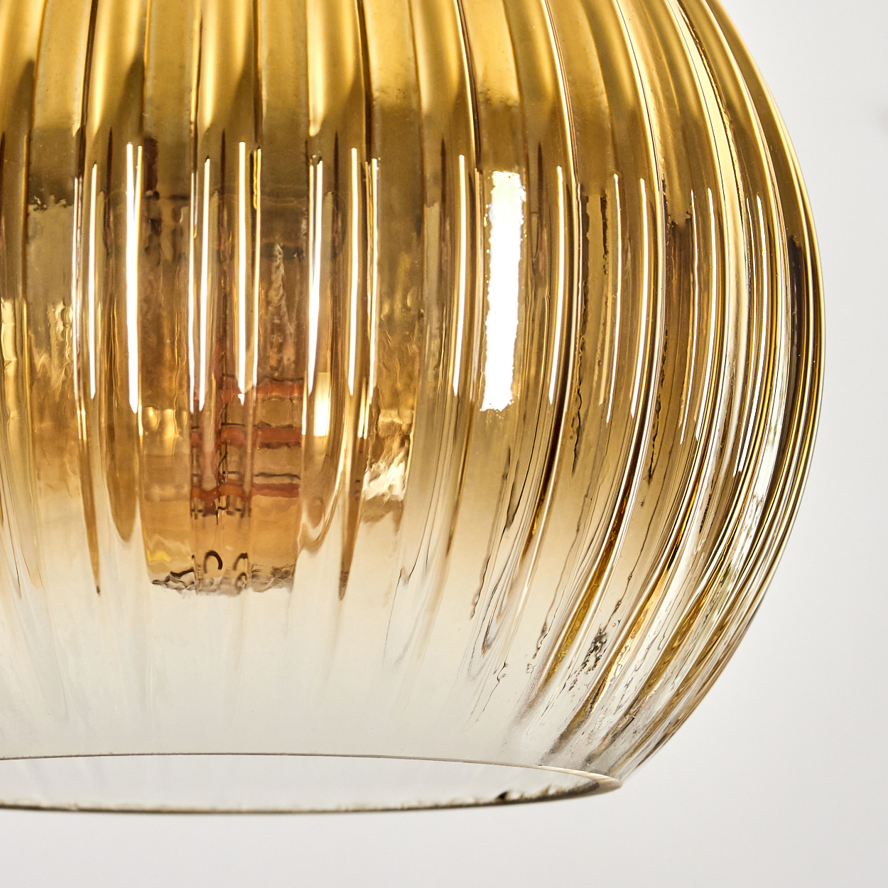Nahaufnahme eines goldenen, gerippten Glaslampenschirms vor weißem Hintergrund. Der Schirm reflektiert Licht, was auf eine interne Glühbirne hindeutet.