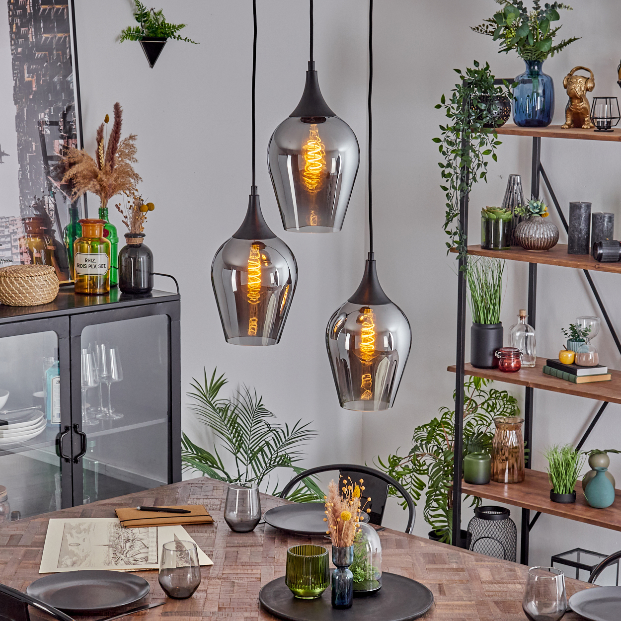 Drei Pendelleuchten hängen über einem Holztisch, der für das Abendessen gedeckt ist. Regale und Schränke mit Dekorationen im Hintergrund.