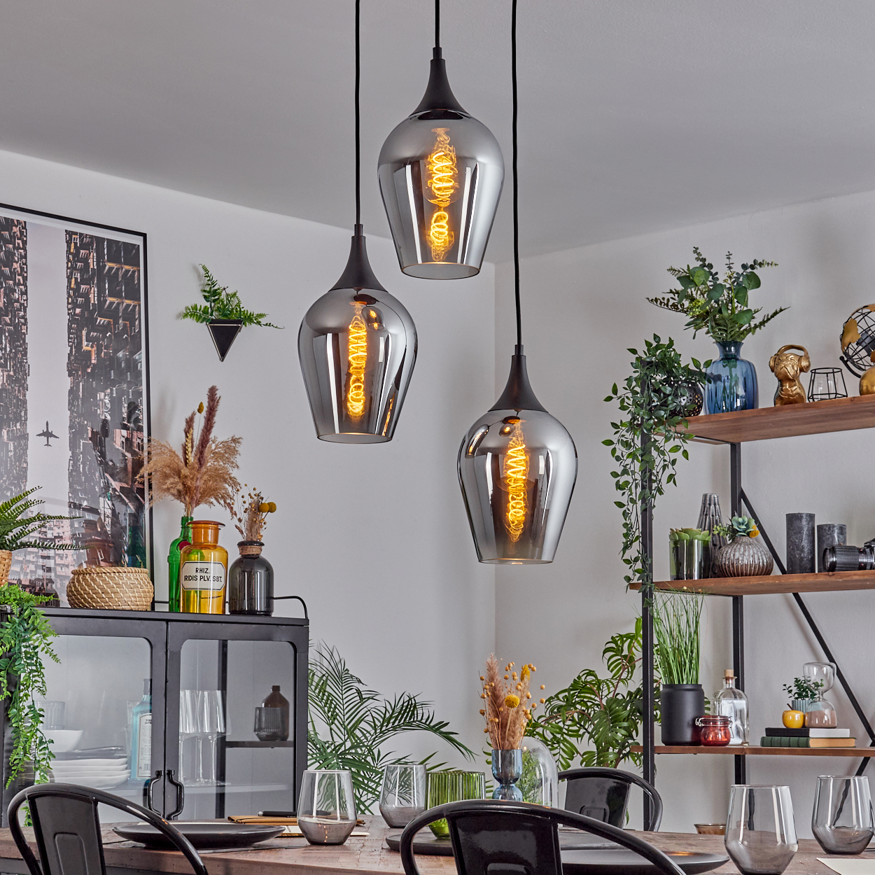 Drei Pendelleuchten mit Edison-Glühbirnen hängen über einem Esstisch. Der Raum hat ein modernes Design mit Pflanzen und Dekoration.