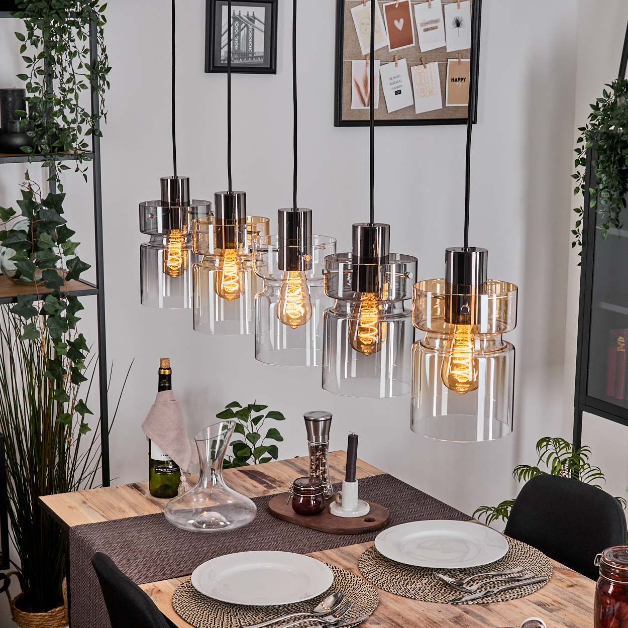 Esszimmer mit Holztisch, Glaslampen, Wein, Karaffe, Tellern und Pflanzen. Lampen haben Edison-Glühbirnen. Schwarze Stühle sichtbar.