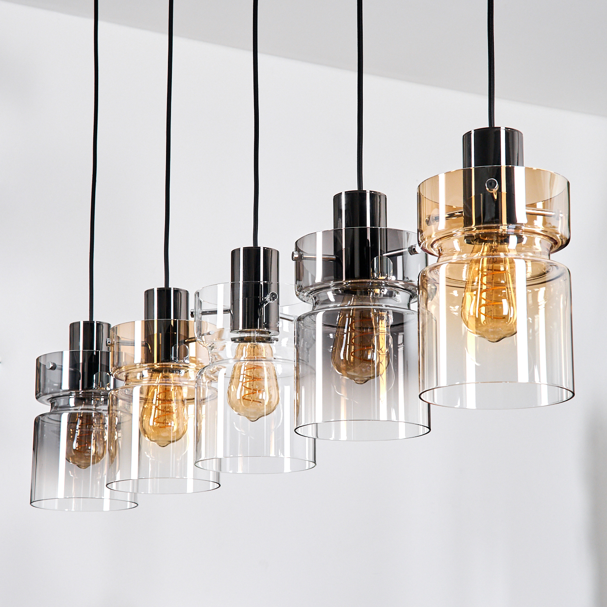 Sechs Hängelampen mit Glasschirmen und Edison-Glühbirnen hängen an schwarzen Kabeln. Die Schirme sind klar und bernsteinfarben, auf weißem Hintergrund.